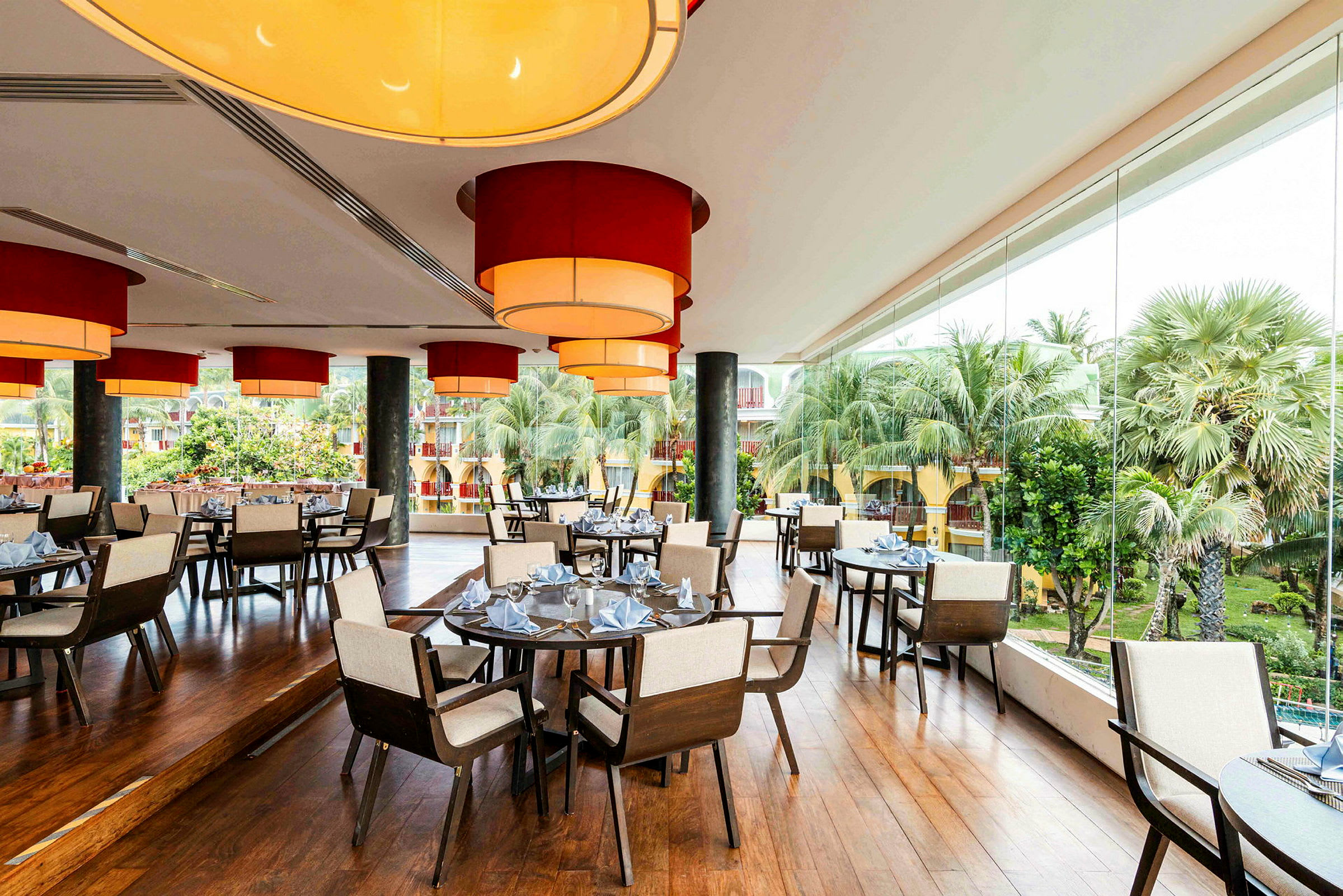 Et indglasset restaurantområde med borde og stole med udsigt over grønne omgivelser og træer. (AI-genereret tekst)