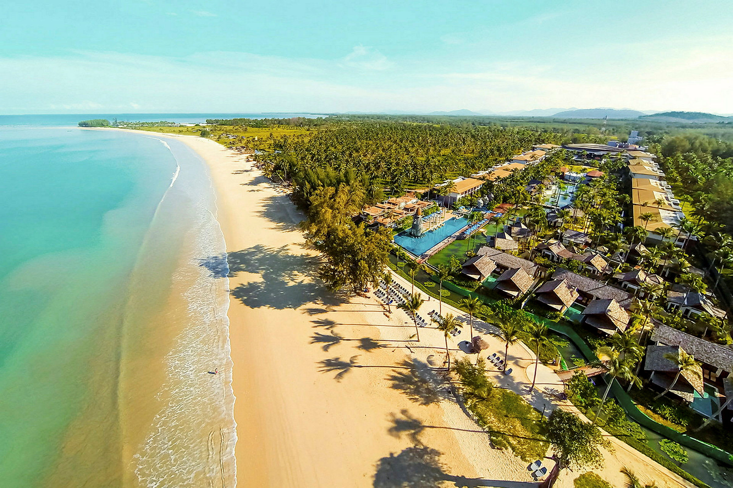 Luftfoto af en lang sandstrand med turkis vand og et feriekompleks med bungalower og pools. (AI-genereret tekst)