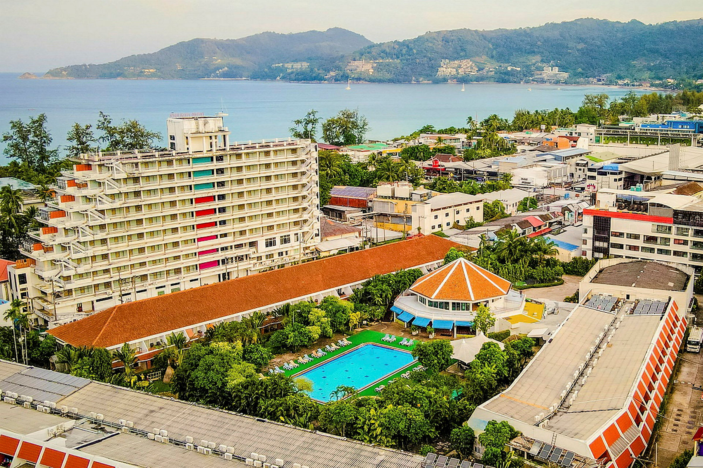 Udsigt over et hotelområde med en hvid bygning i flere etager, en swimmingpool og udsigt mod havet. (AI-genereret tekst)