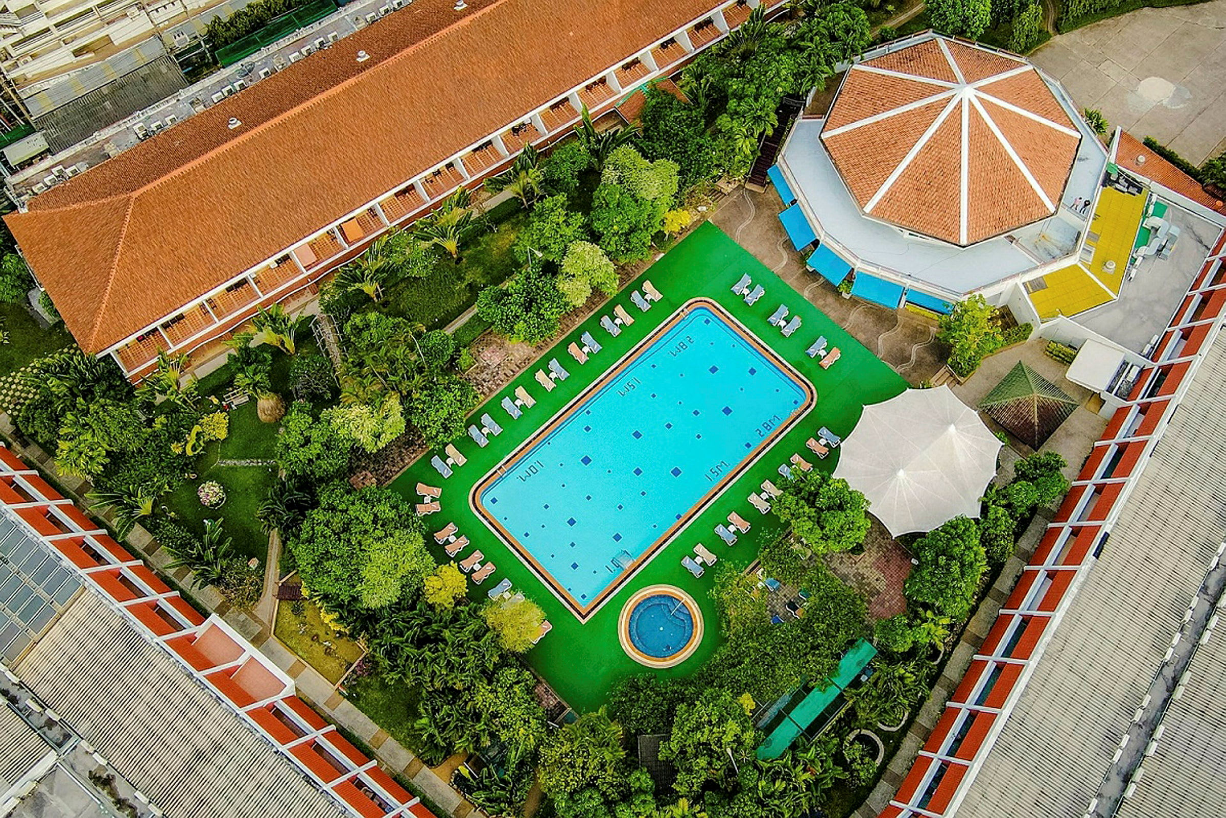 Luftfoto af et hotelområde med en pool omgivet af liggestole samt frodig vegetation og bygninger. (AI-genereret tekst)