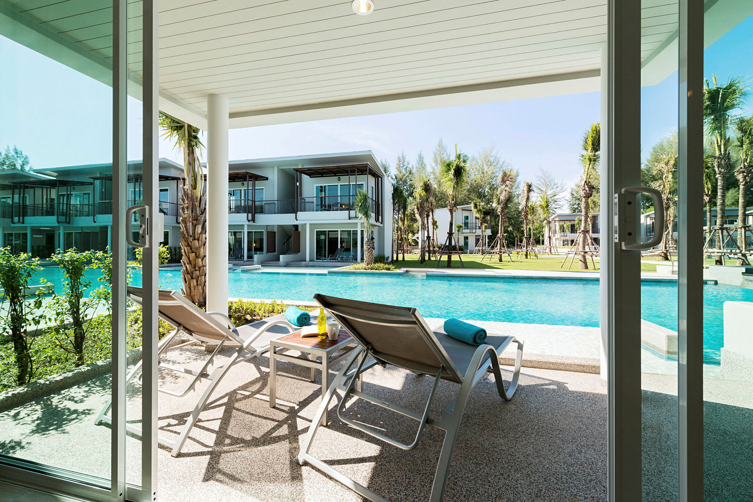 Billede af en terrasse med liggestole og et bord, ved en pool med moderne bygninger i baggrunden. (AI-genereret tekst)