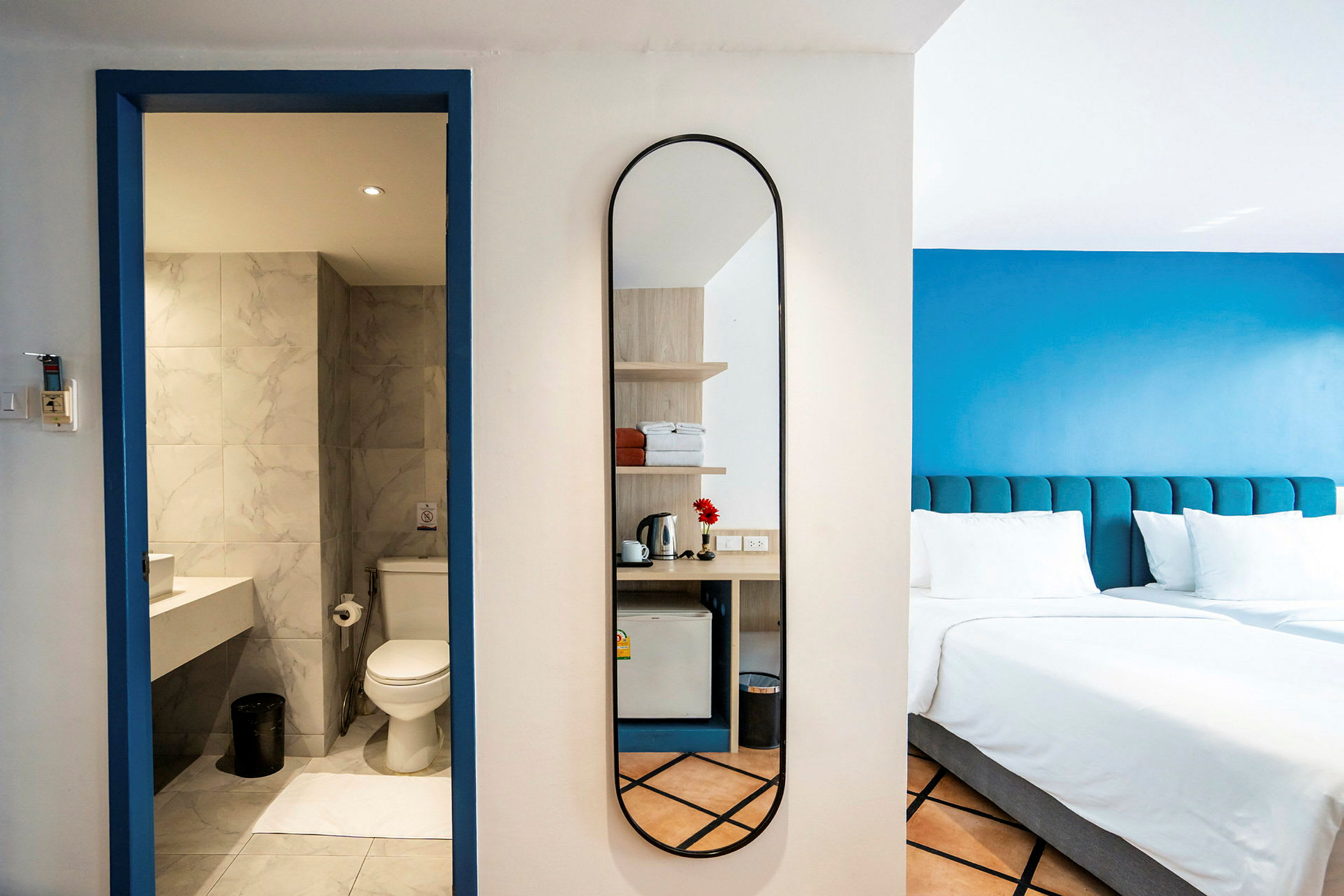 Et hotelværelse med et spejl, et badeværelse med toilet og håndvask samt en seng med blå sengegavl. (AI-genereret tekst)
