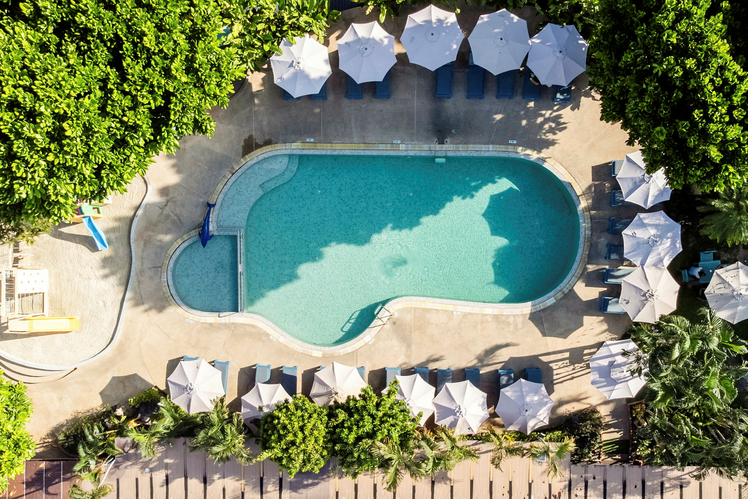 En oval pool omgivet af liggestole og parasoller samt grønne områder. (AI-genereret tekst)