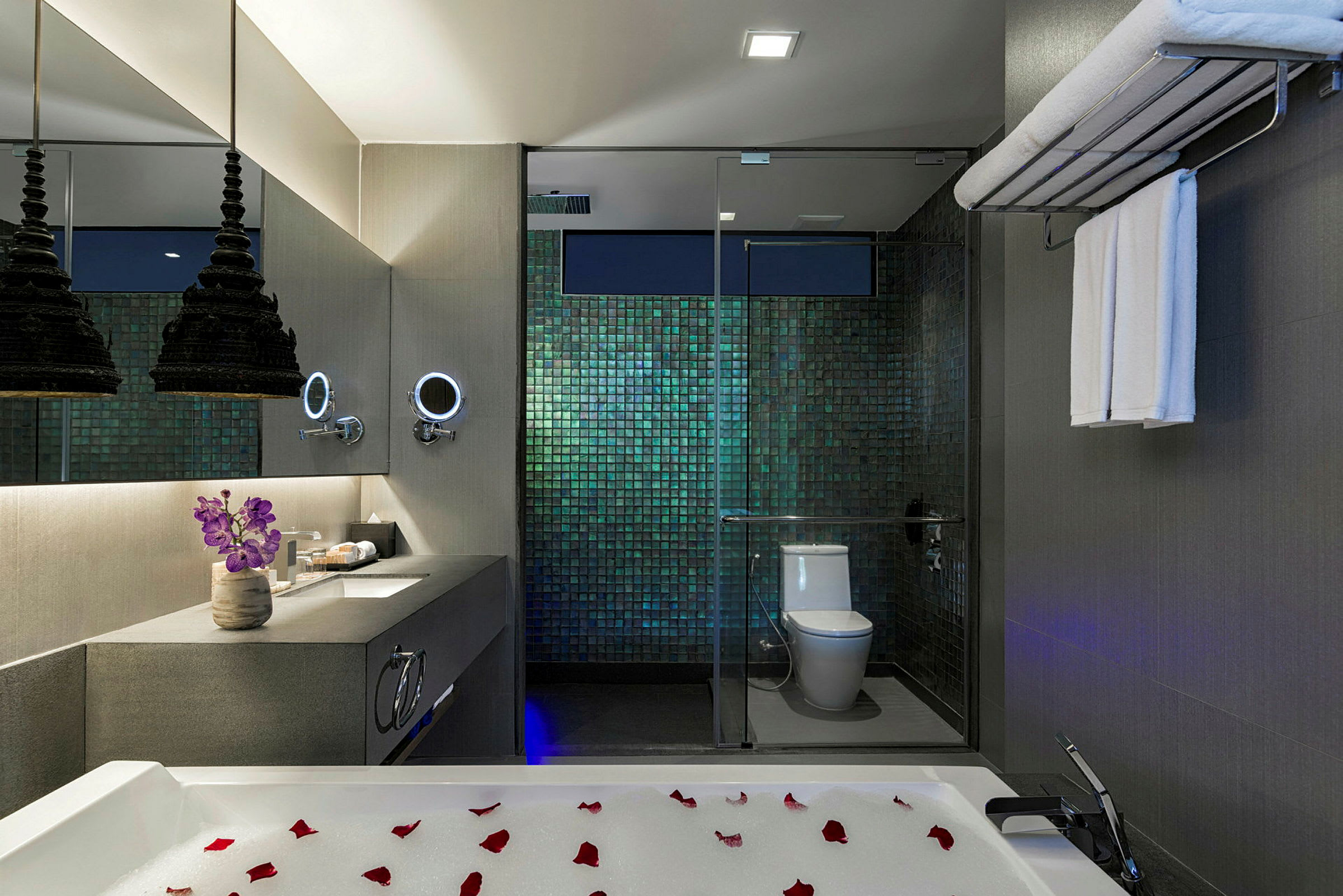 Et moderne badeværelse med luksuriøst design. Der er et badekar med skumbad og rosenblade i forgrunden. En elegant håndvask med spejl, blomster og belysning samt en bruser og toilet bag glasvægge kan ses. (AI-genereret tekst)