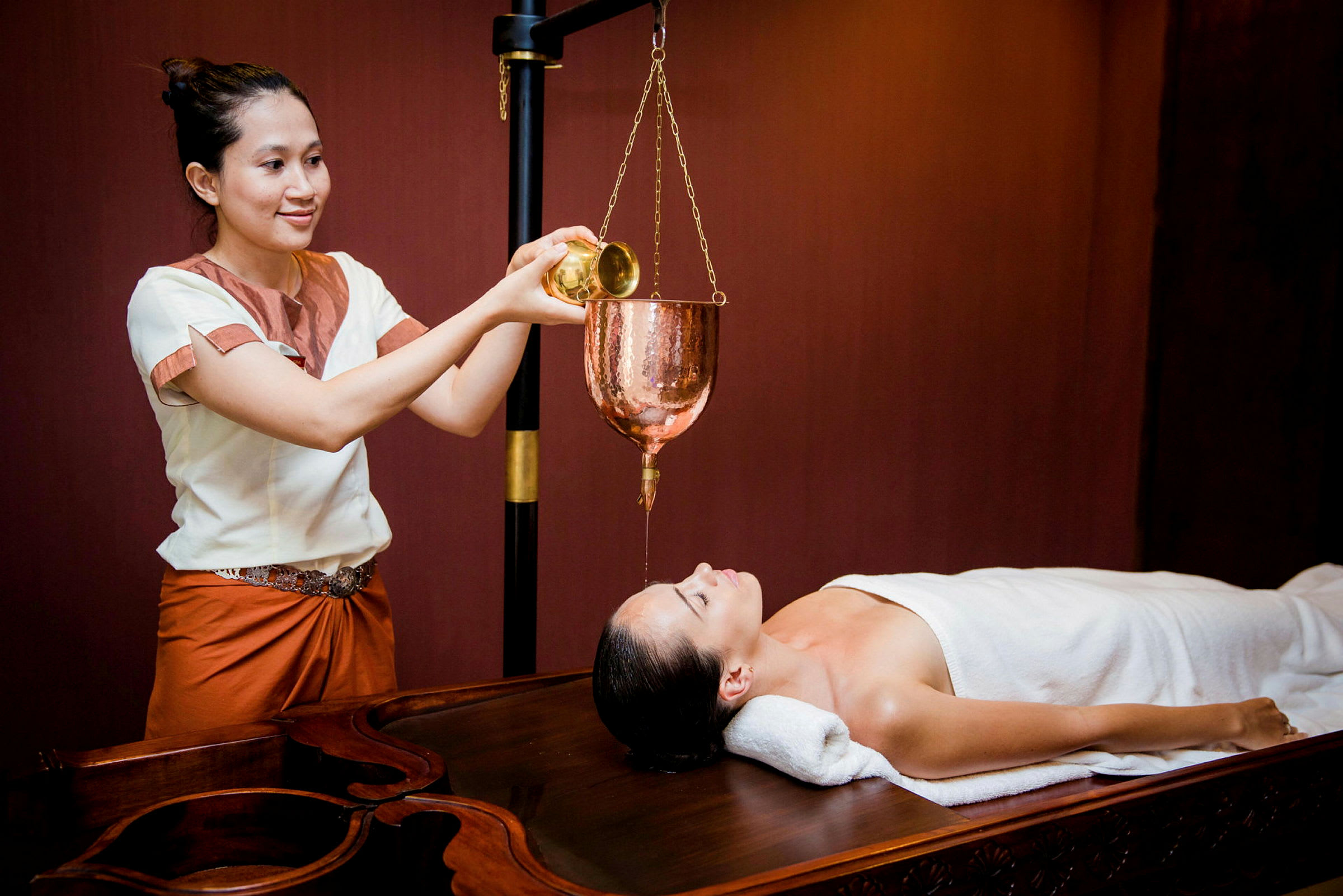 En person udfører en spa-behandling med olie, der hældes fra en kobberbeholder på en person liggende på en massagebriks. (AI-genereret tekst)