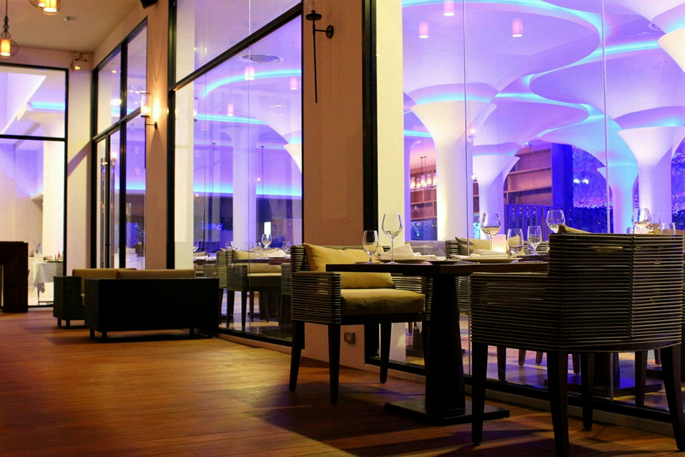 Interiør af en elegant restaurant med glasvægge og moderne møbler, belysning i blåt og varmt lys. (AI-genereret tekst)