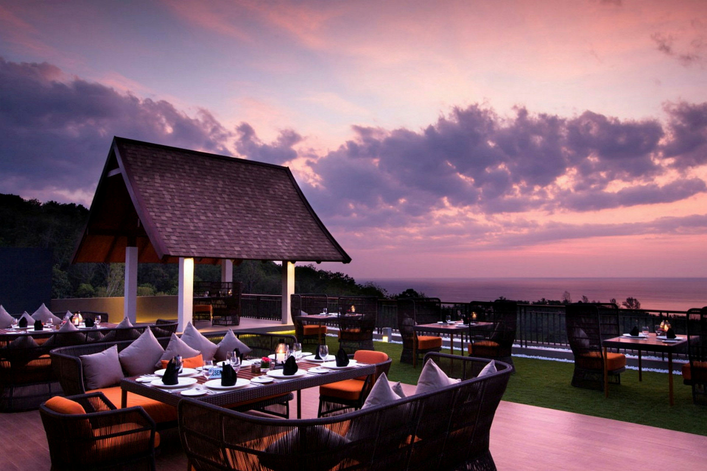 En udendørsrestaurant med borde og stole under en lilla og lyserød himmel ved solnedgang, med udsigt over havet. (AI-genereret tekst)