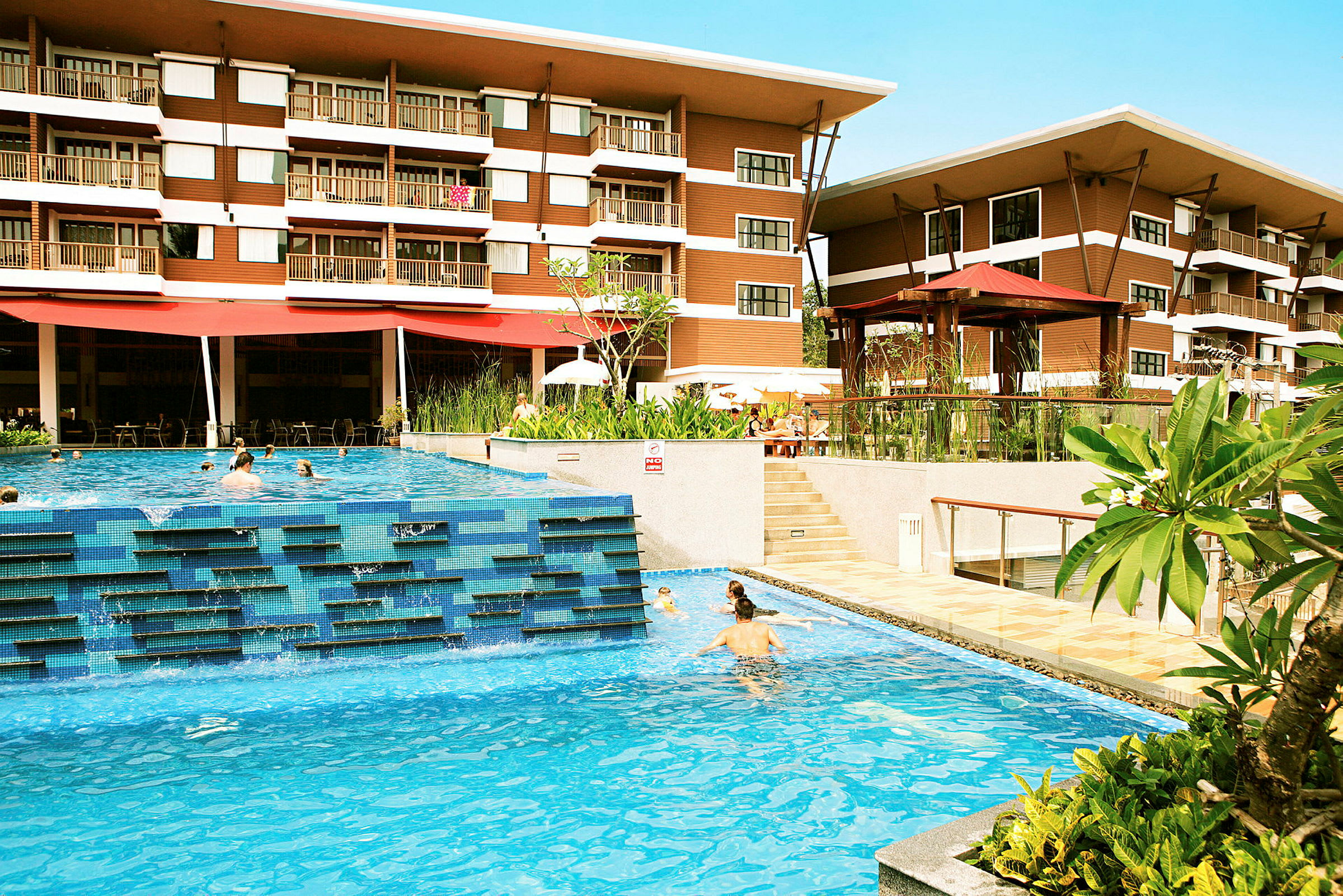 Et hotel med to etager er synligt bag en udendørs pool, hvor mennesker svømmer. Grønne planter og terrasseområder omkranser poolområdet. (AI-genereret tekst)