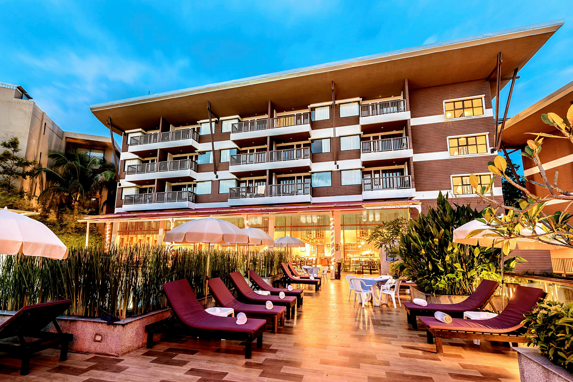 Billede af et hotel med flere etager i aftenbelysning. Foran hotellet er der liggestole og parasoller. (AI-genereret tekst)