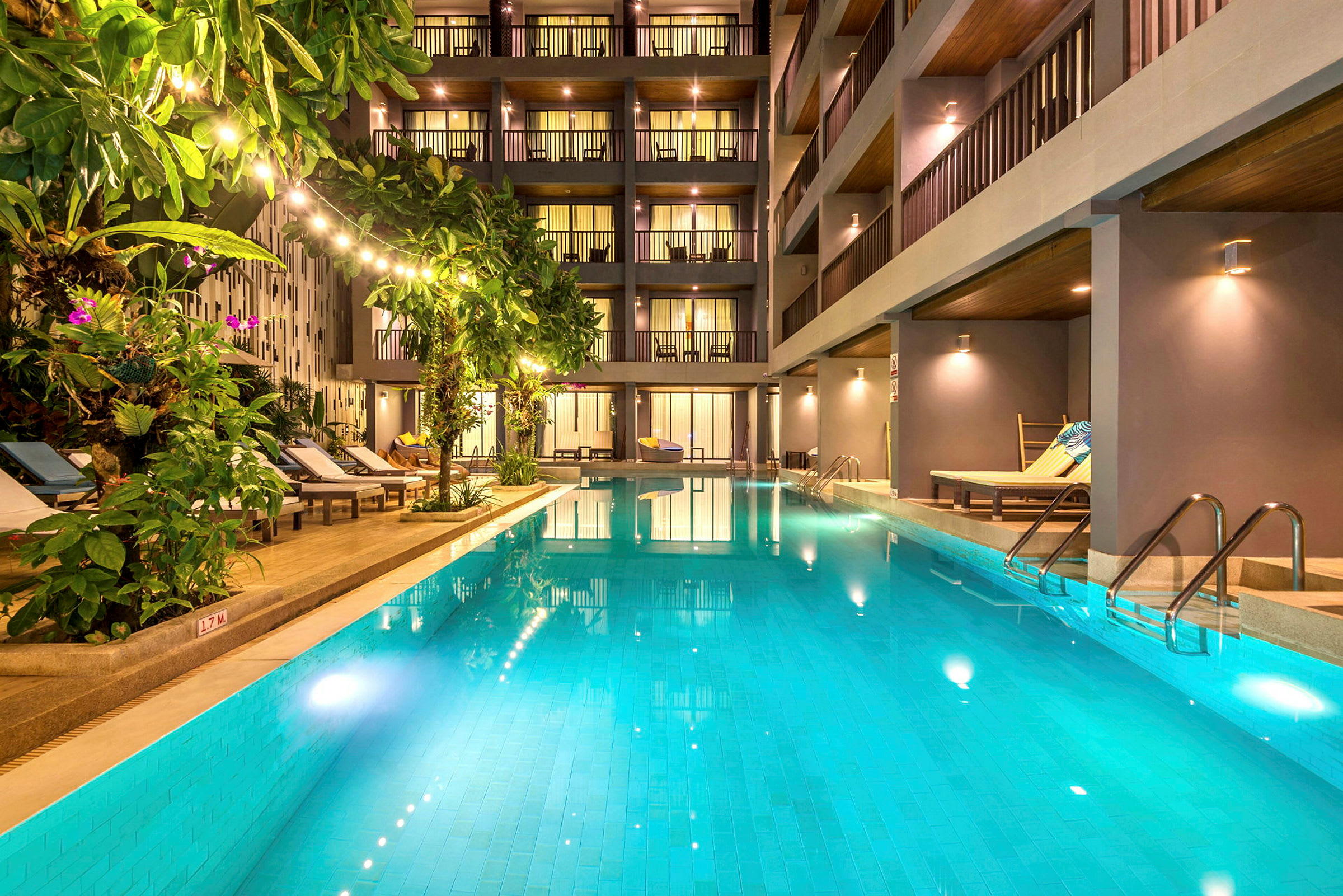 En udendørs pool omgivet af liggestole og grønne planter foran et moderne hotel. (AI-genereret tekst)