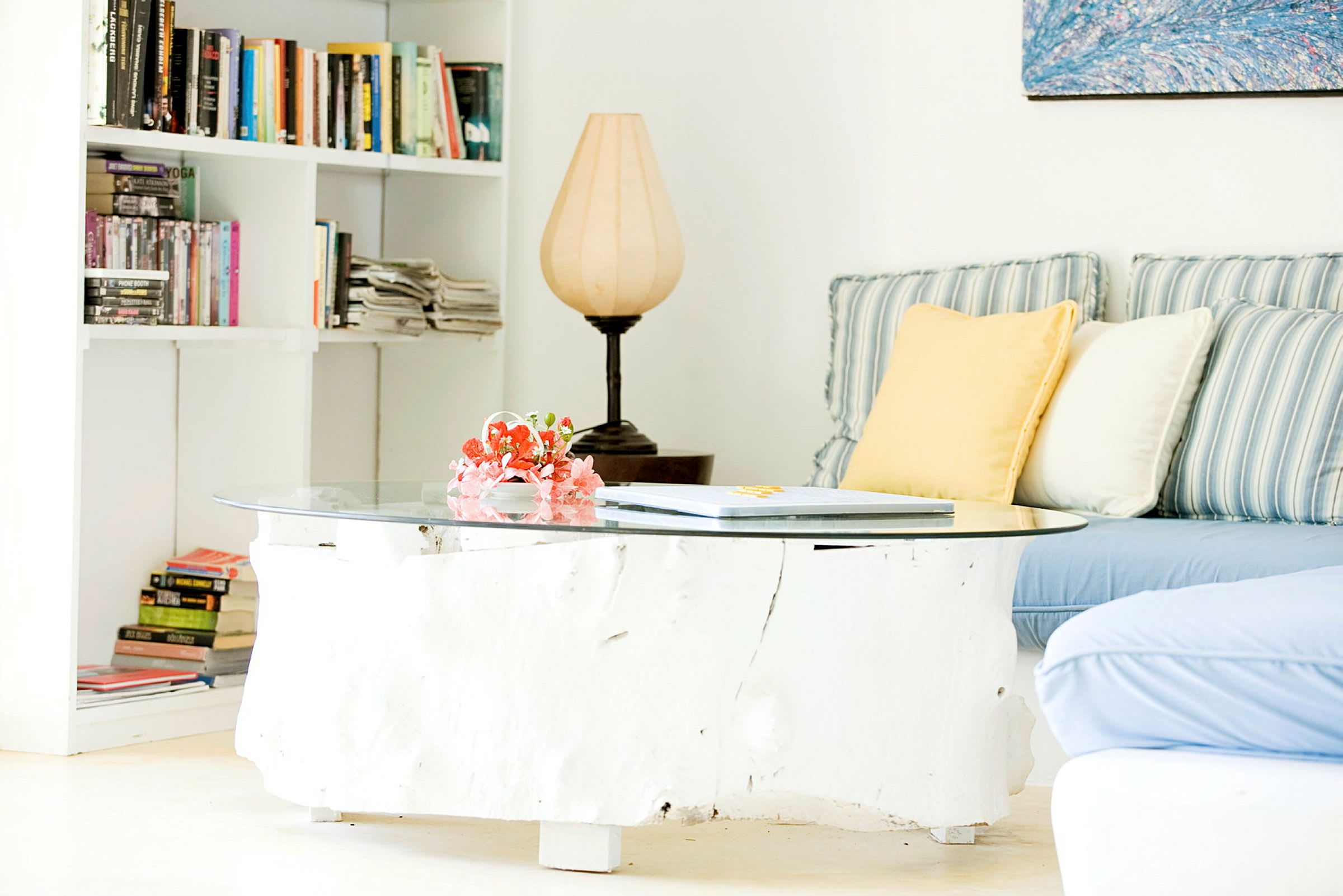 En stue med en sofa med stribede og ensfarvede puder. Et sofabord i træ med en glasplade øverst, dekoreret med blomster. En bogreol fyldt med bøger og aviser samt en lampe ses i baggrunden. (AI-genereret tekst)