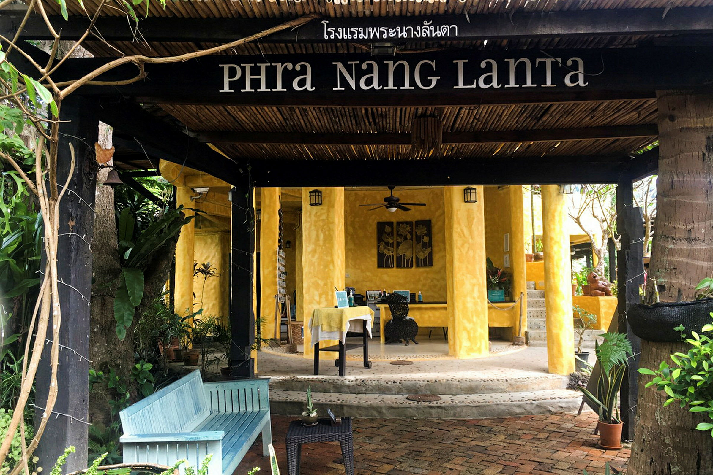 Indgangen til et hotel med navnet Phra Nang Lanta, med gule vægge og trædetaljer, omgivet af grønt. (AI-genereret tekst)