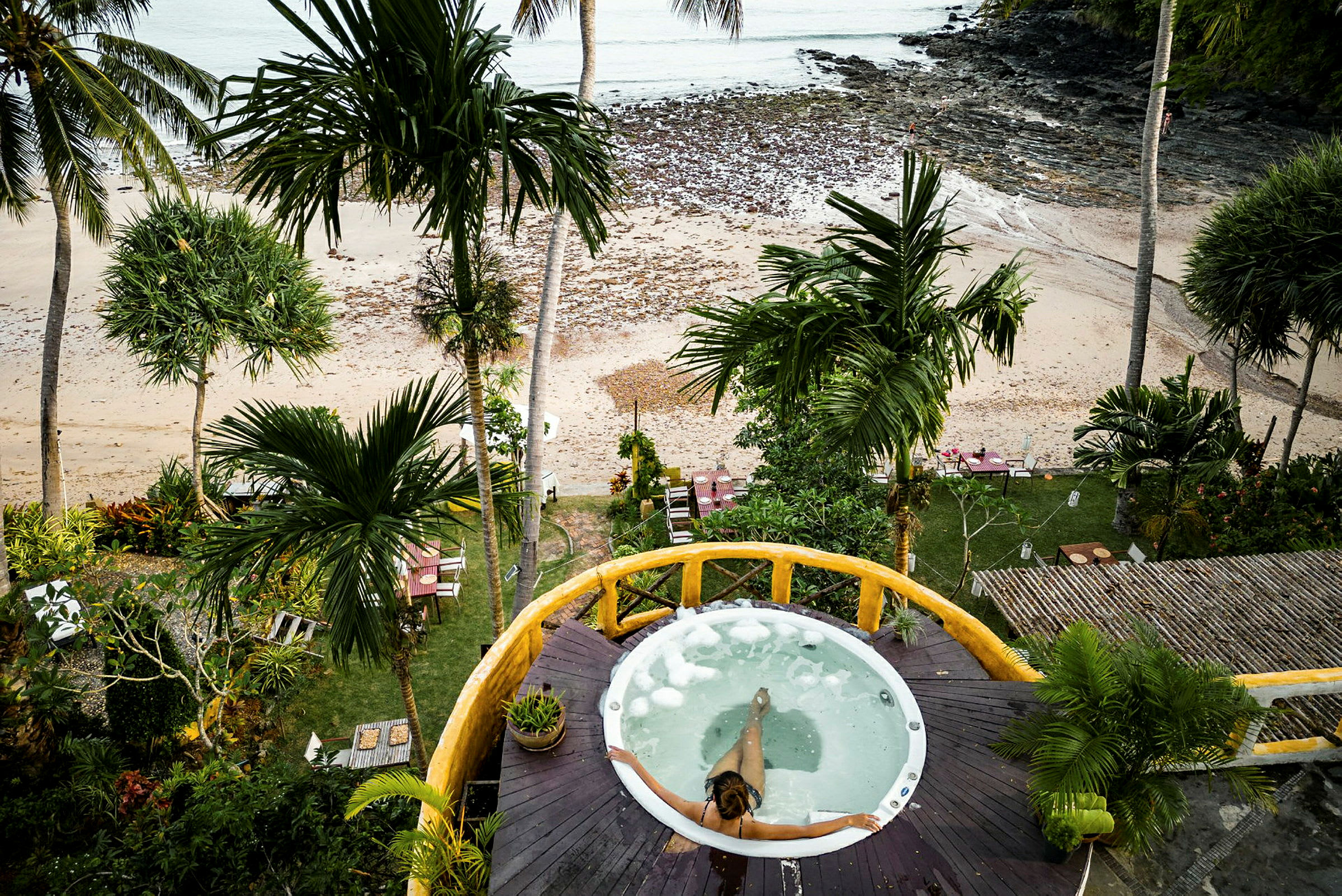En person sidder i en rund jacuzzi på en udendørs terrasse med udsigt over en tropisk strand. (AI-genereret tekst)