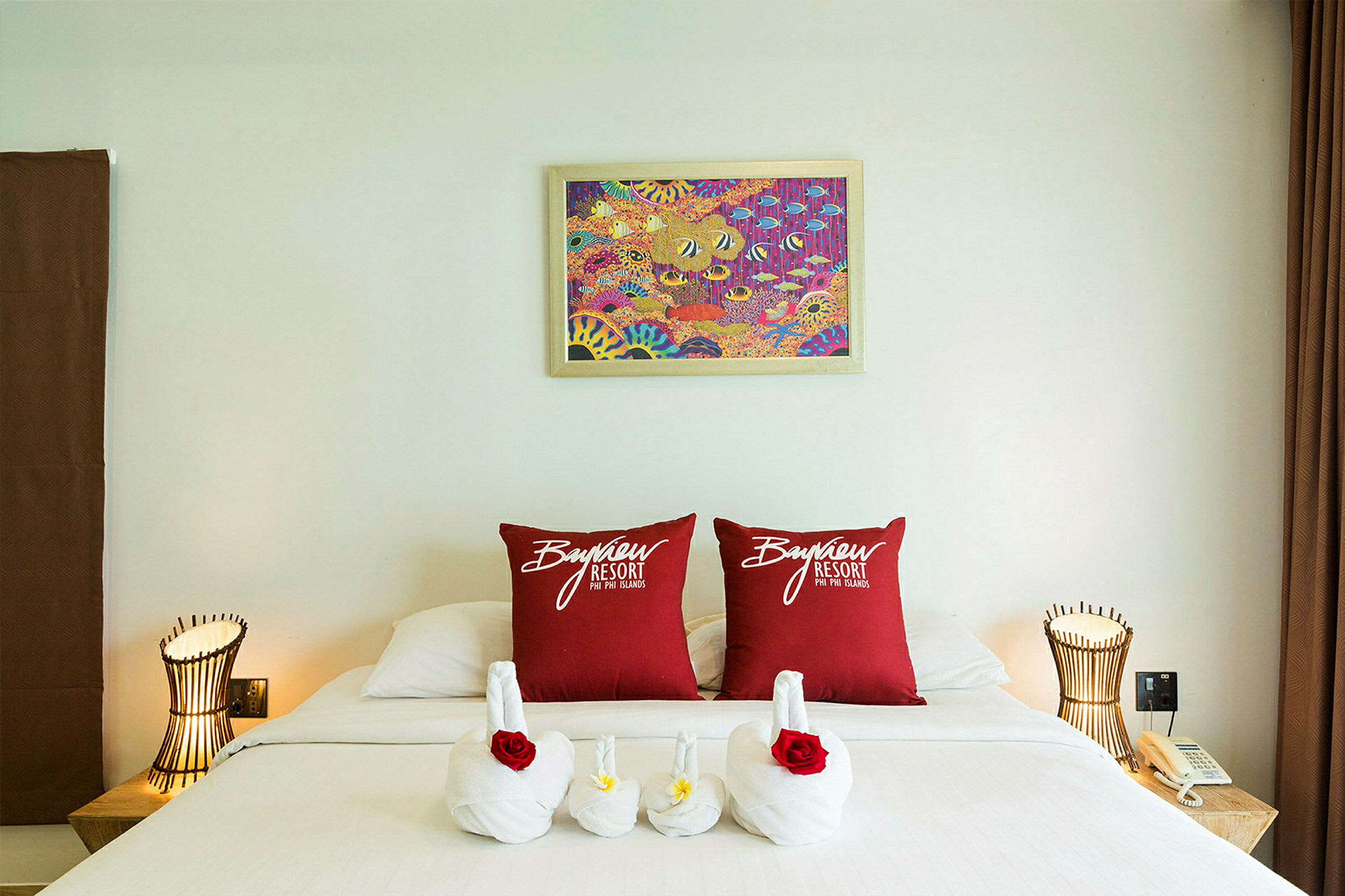 Et hotelværelse med en dekoreret seng. To røde puder med teksten "Bayview Resort" er placeret på sengen, og håndklæder er arrangeret som svaner med blomster. Der er en lampe og en telefon på et natbord. (AI-genereret tekst)