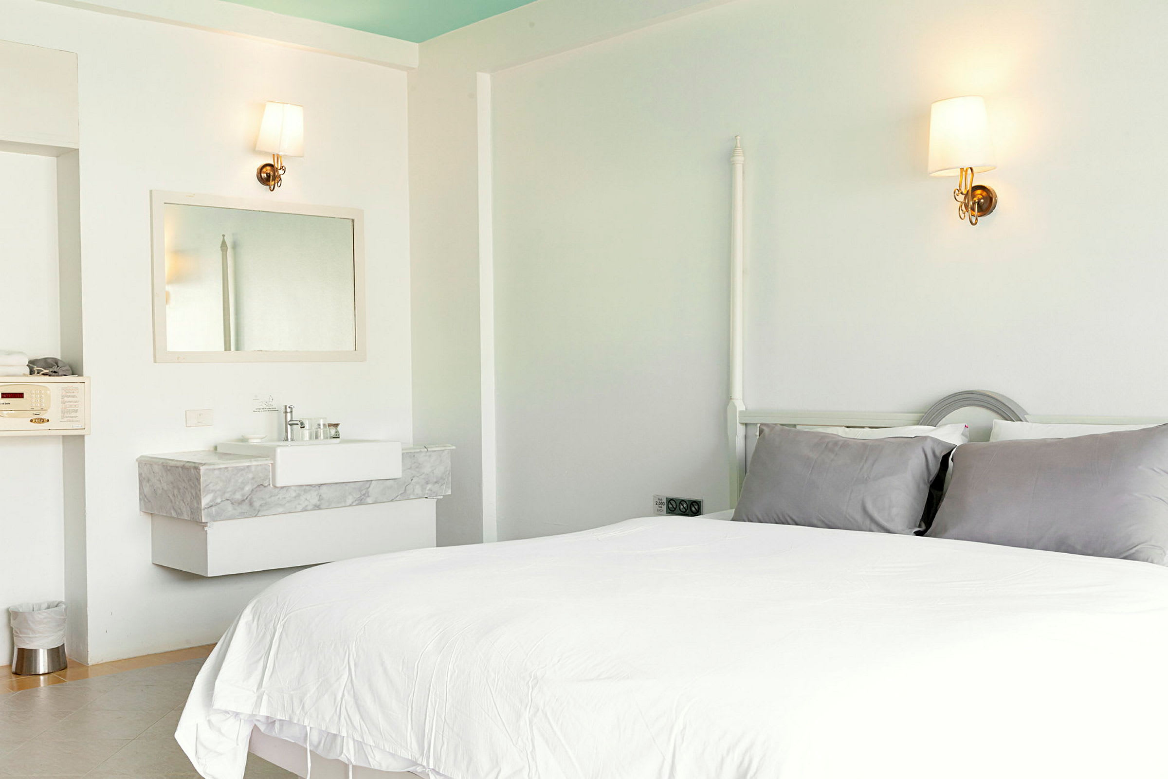 Et lyst hotelværelse med en seng, grå puder og en håndvask med et spejl. (AI-genereret tekst)