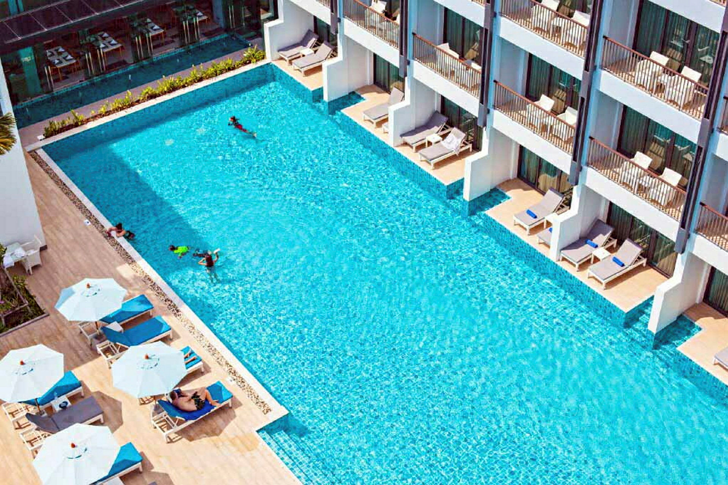 En udendørs pool med liggestole og parasoller ved siden af samt bygninger med terrasser. (AI-genereret tekst)