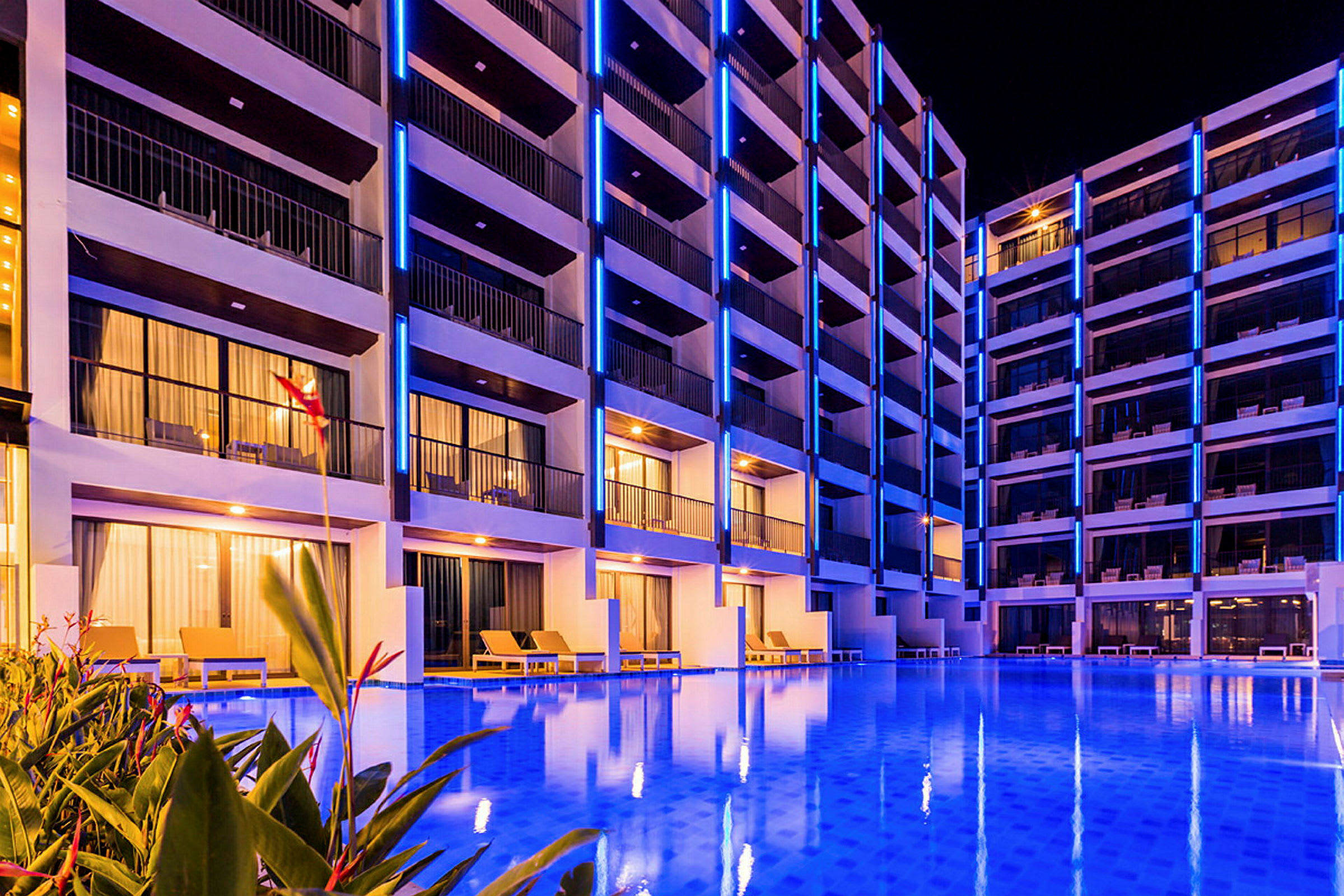 Et billede af et hotel med flere etager, oplyst med blå neonlys og en pool i forgrunden. (AI-genereret tekst)