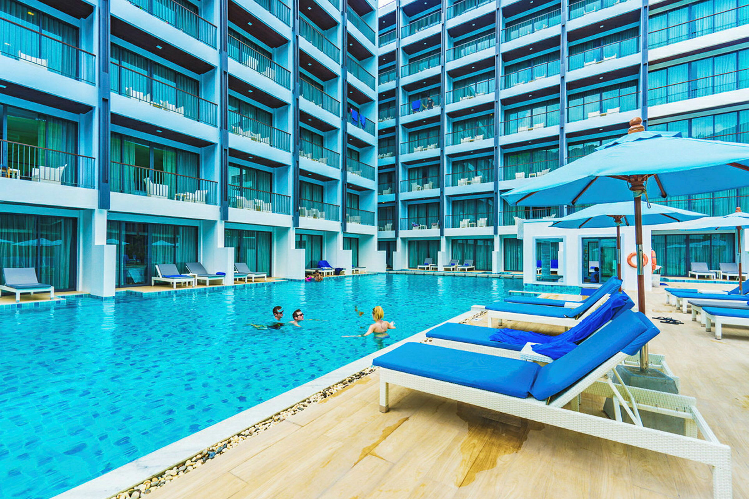 En stor pool omgivet af et moderne fleretagers hotel, med liggestole og parasoller ved kanten. (AI-genereret tekst)
