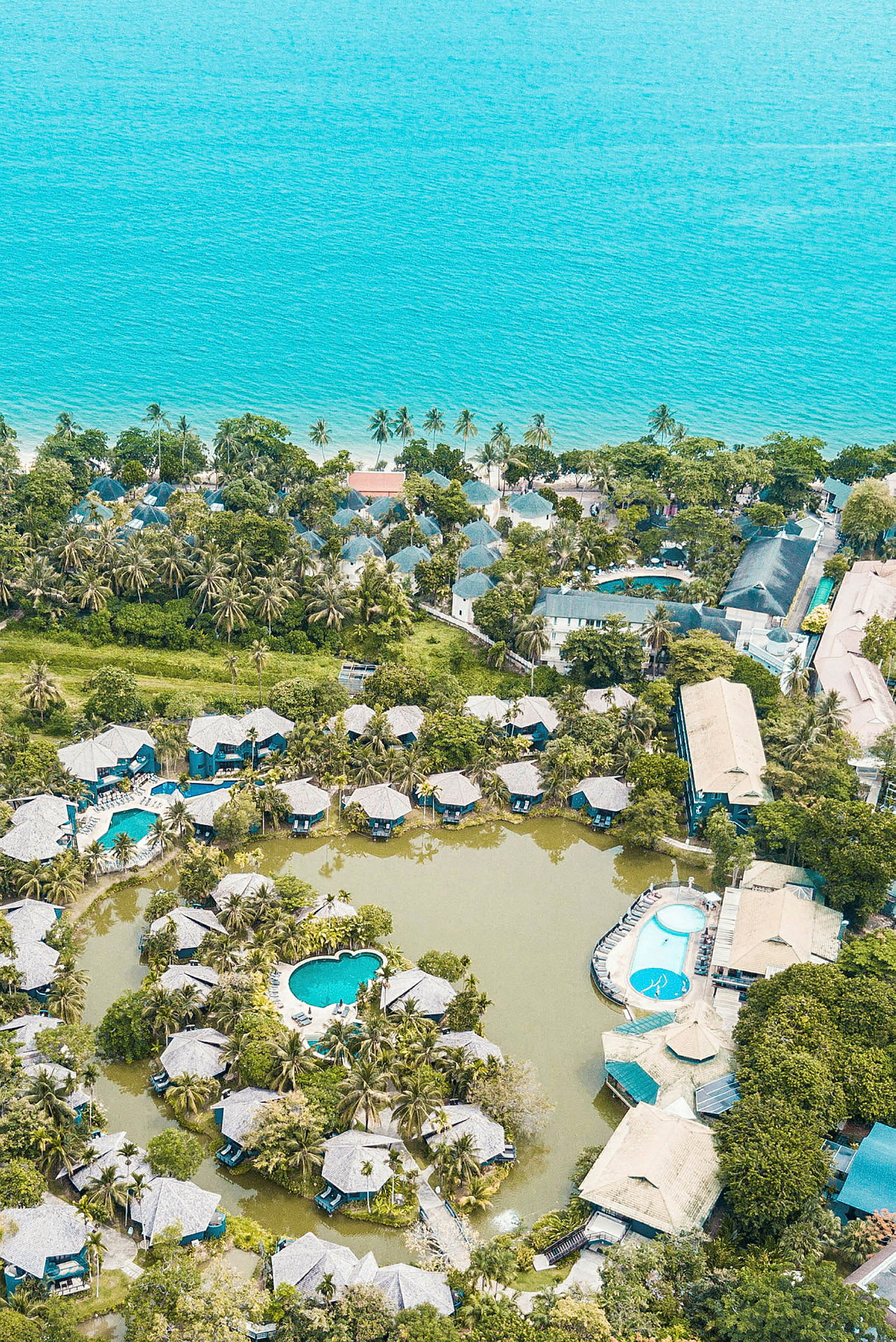 Luftfoto af en resort ved havet med bungalower omkring pools og grønne områder. (AI-genereret tekst)