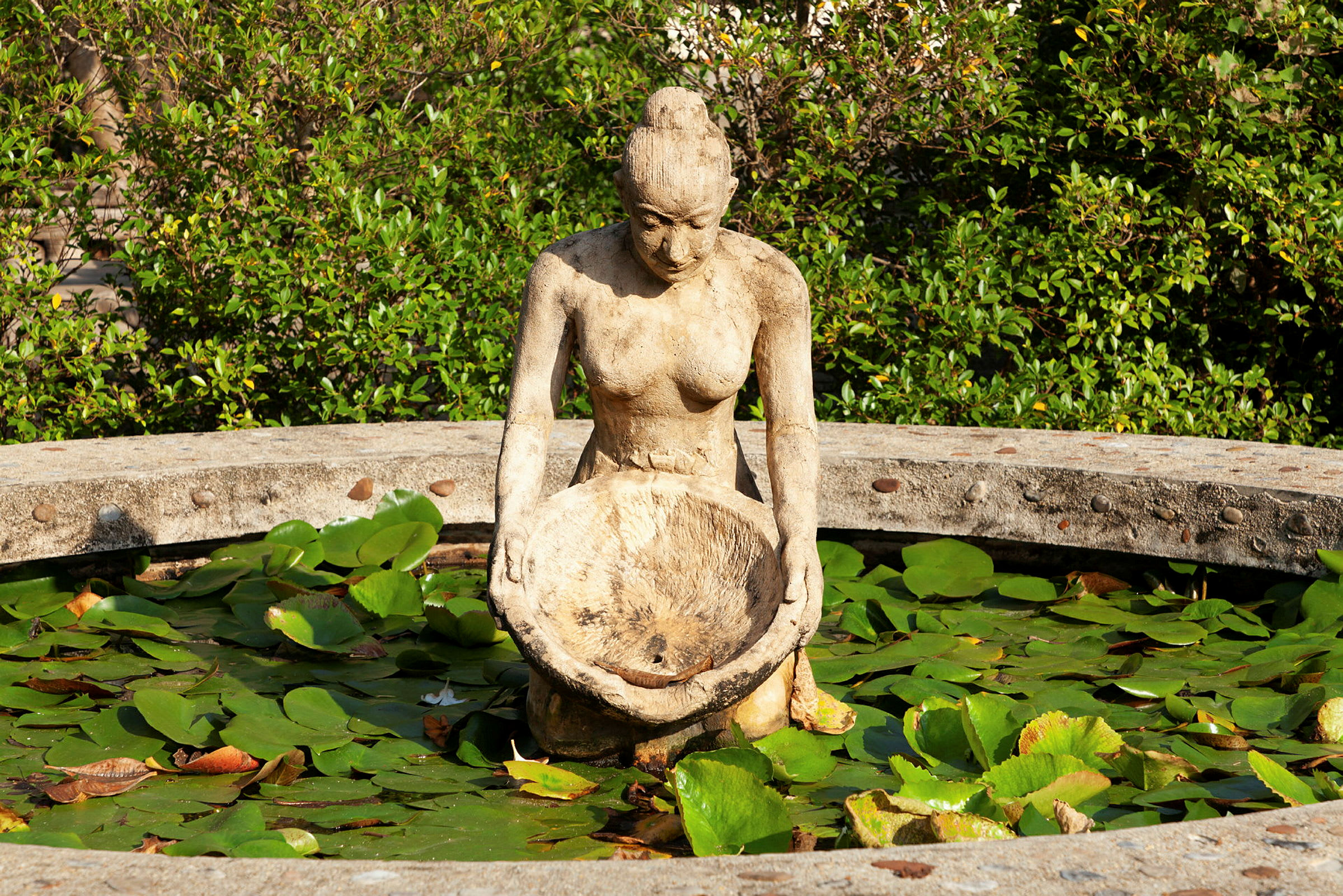 Statue af en kvinde, der holder en skål, omgivet af et cirkulært vandfyldt kar med åkandeblade. (AI-genereret tekst)