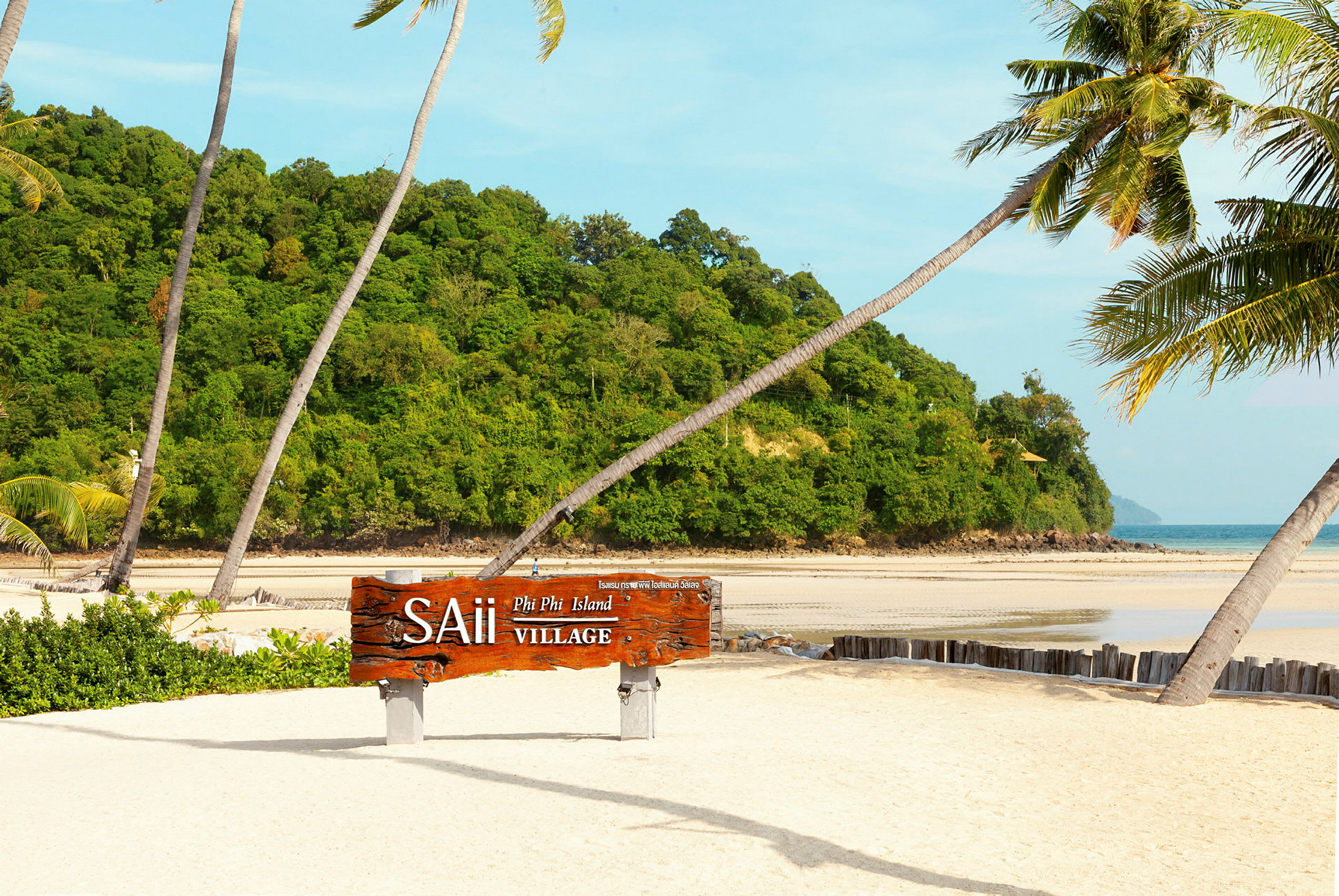 Sandstrand med palmer og et træskilt med teksten "SAii Phi Phi Island Village", omgivet af grøn vegetation og havet i baggrunden. (AI-genereret tekst)