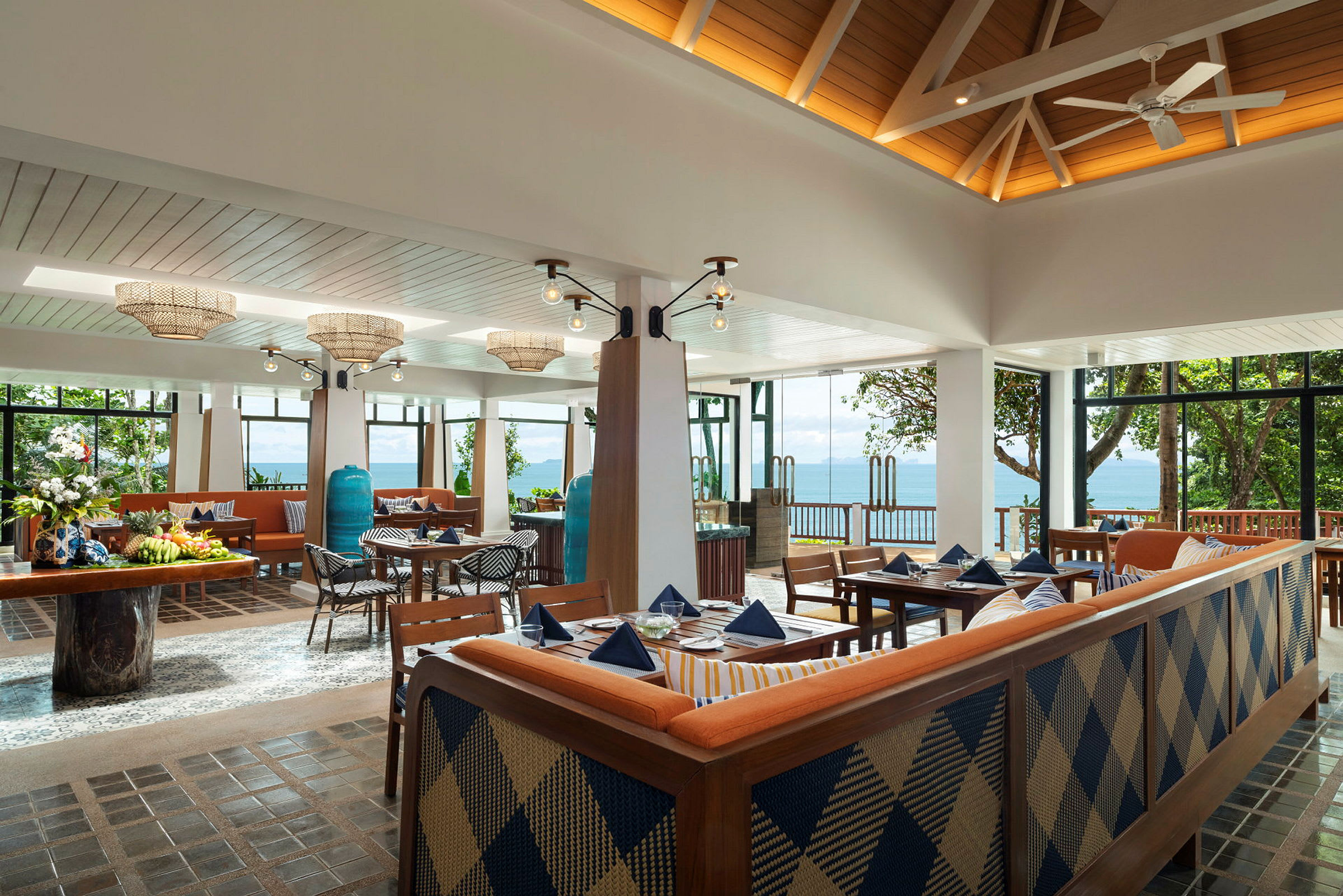 Et luftigt og lyst restaurantområde med store vinduer ud mod havet. Møblerne består af borde med blå duge, træstole og sofaer. (AI-genereret tekst)