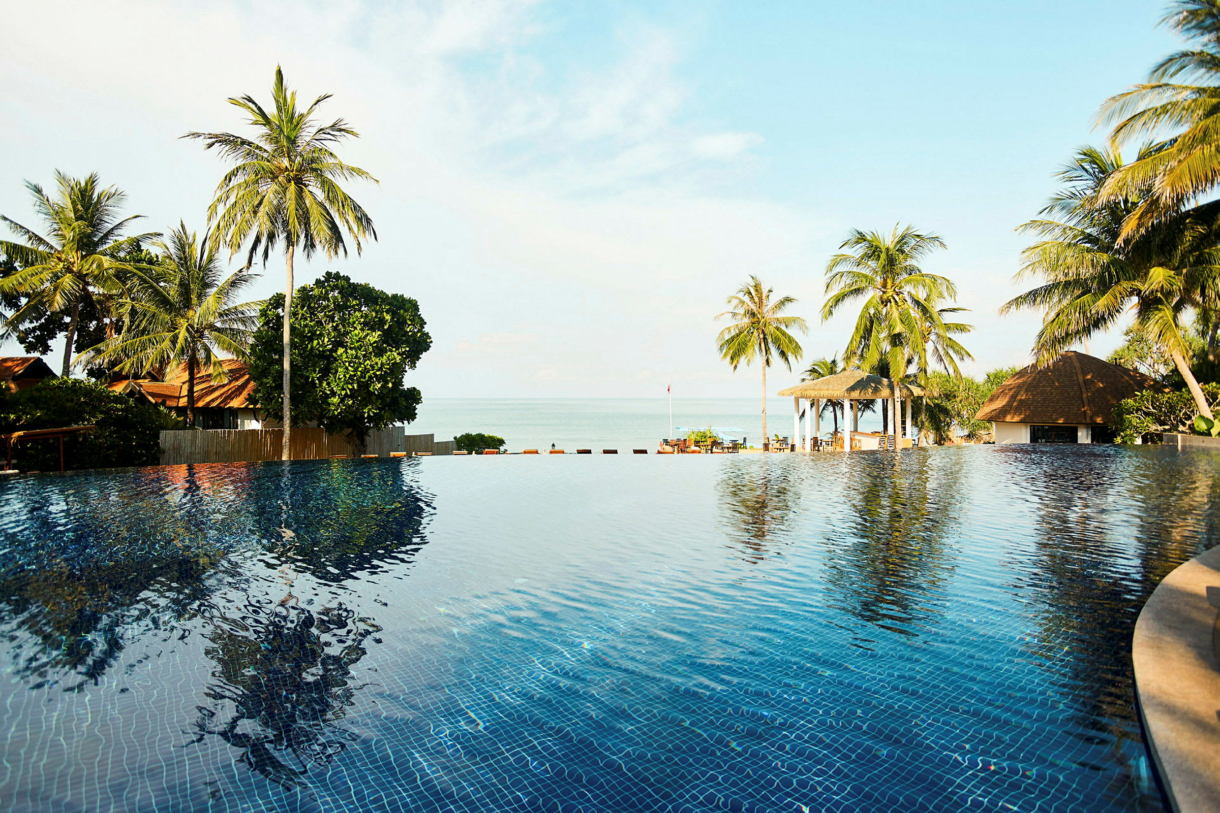 En infinitypool med spejlblankt vand, omgivet af palmer og bygninger med bungalows tæt på havet. (AI-genereret tekst)