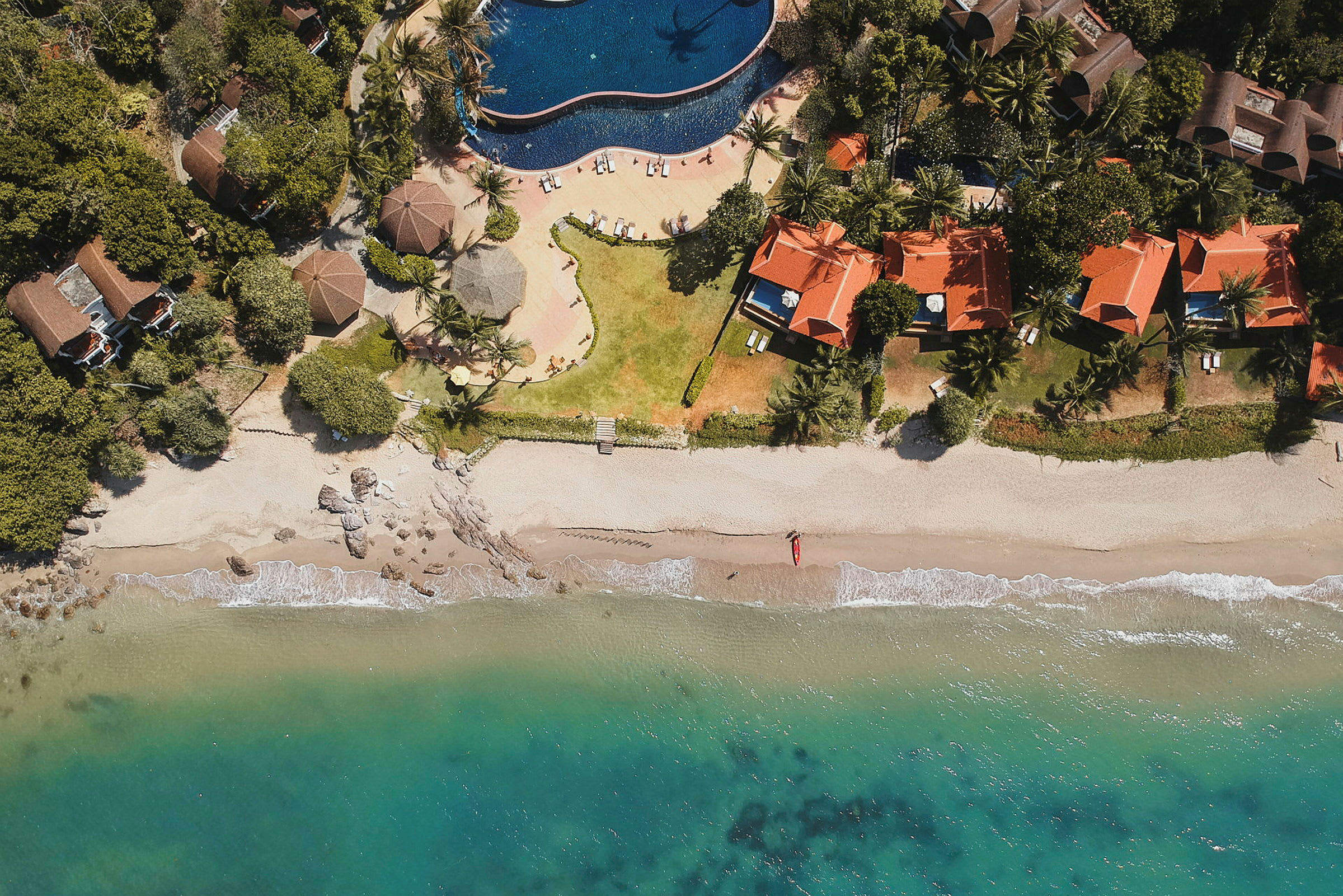 Luftfoto af en strand med turkisblåt vand, sand, hytter med røde tage og en swimmingpool. (AI-genereret tekst)