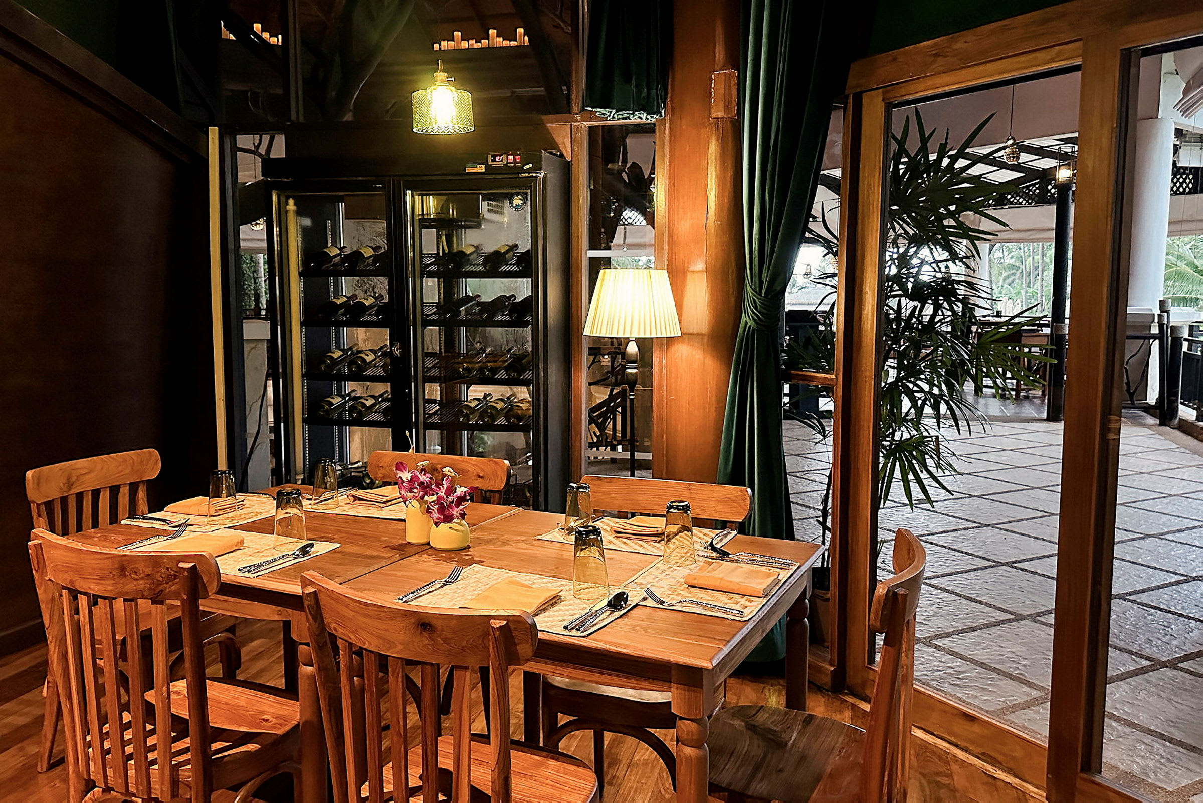 Et træbord med træstole er dækket til fire personer i en restaurantmiljø. Vinhylder og grønne gardiner er synlige i baggrunden. (AI-genereret tekst)