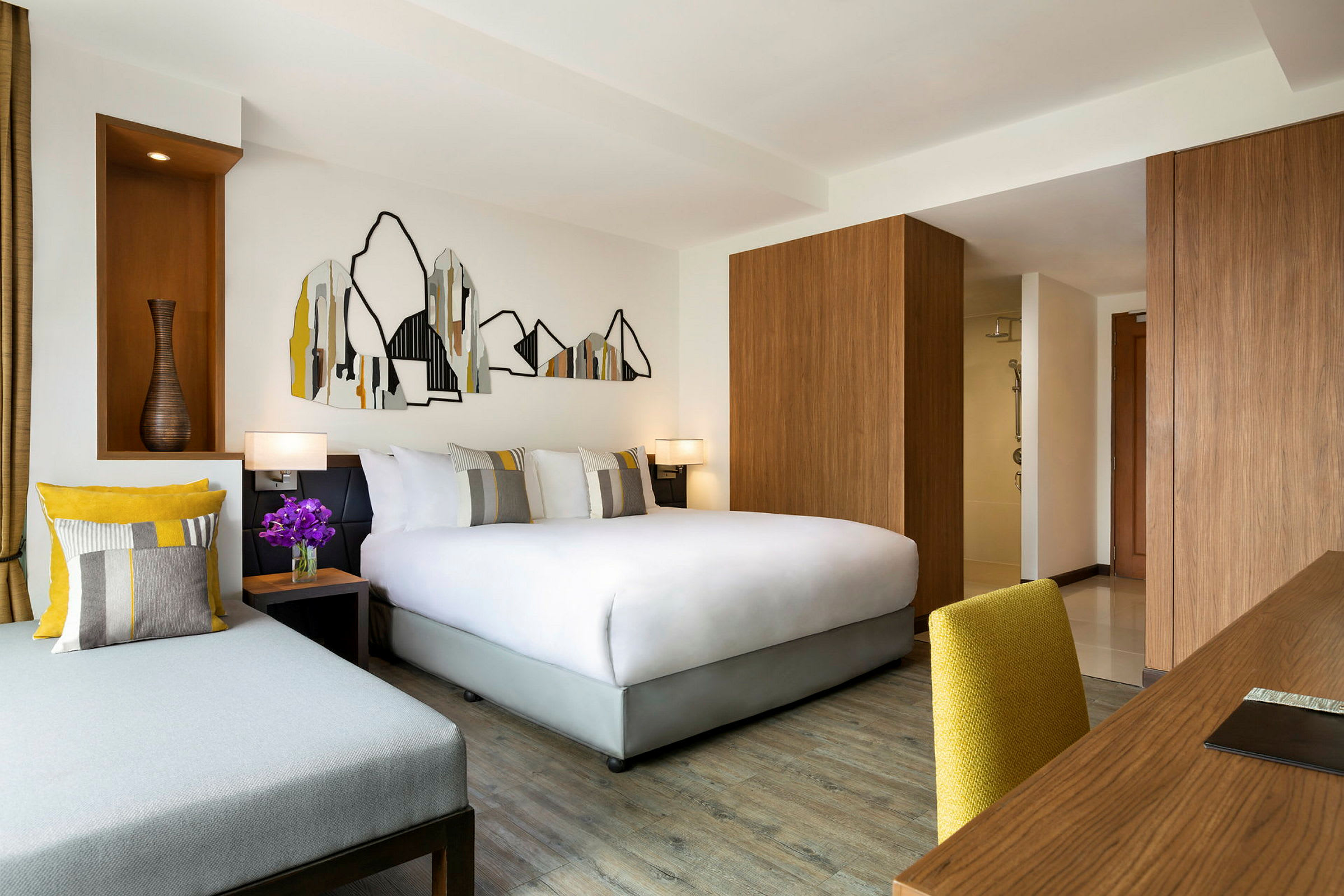 Et moderne hotelværelse med en dobbeltseng, en sofa og et skrivebord med en gul stol. Væggdekorationer med bjergmotiv i abstrakt stil. (AI-genereret tekst)