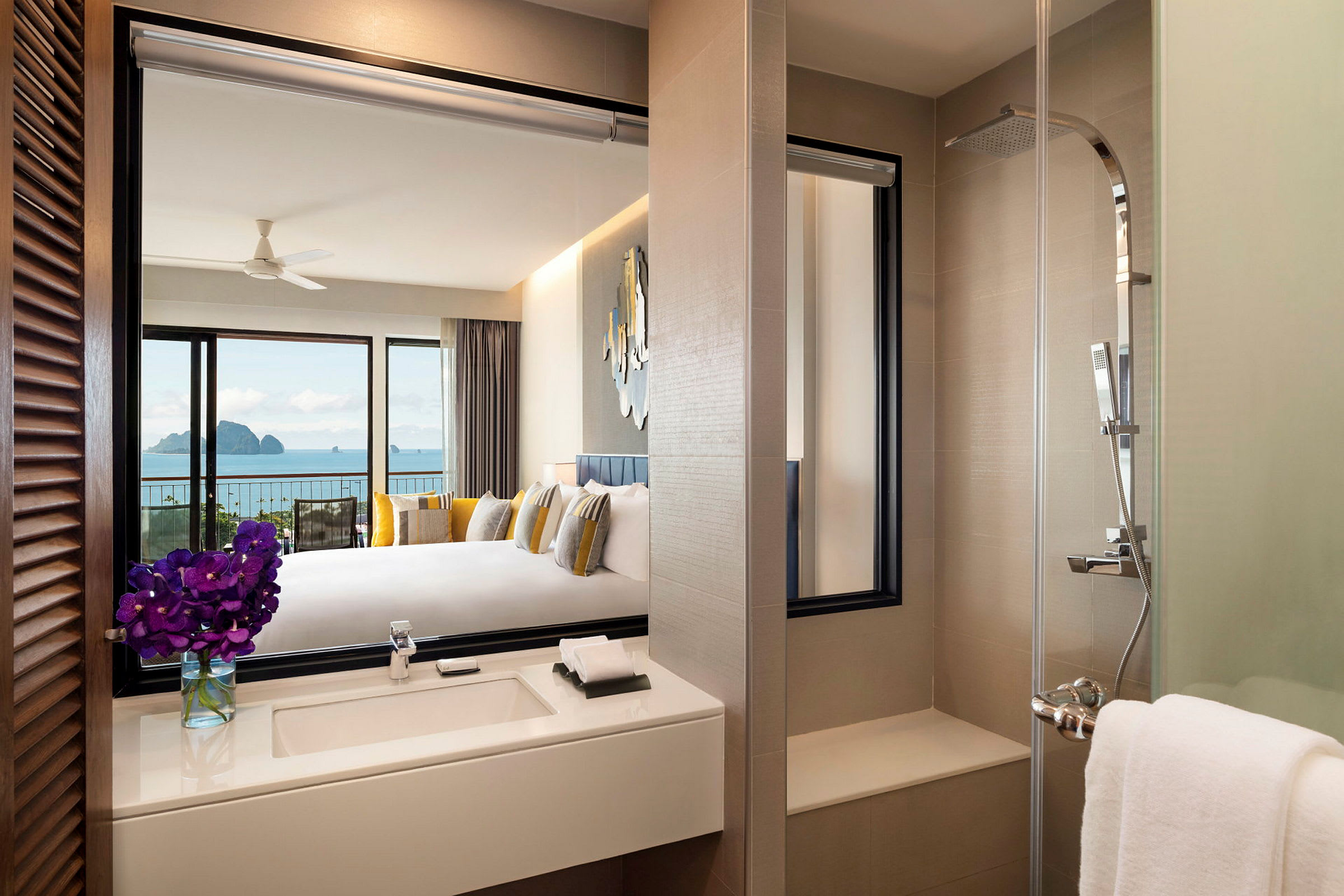 Et moderne hotelværelse med udsigt over havet. Billedet viser badeværelset med en håndvask med blomster, samt en bruser i forgrunden og et tilstødende soveværelse i baggrunden med stor seng og havudsigt. (AI-genereret tekst)