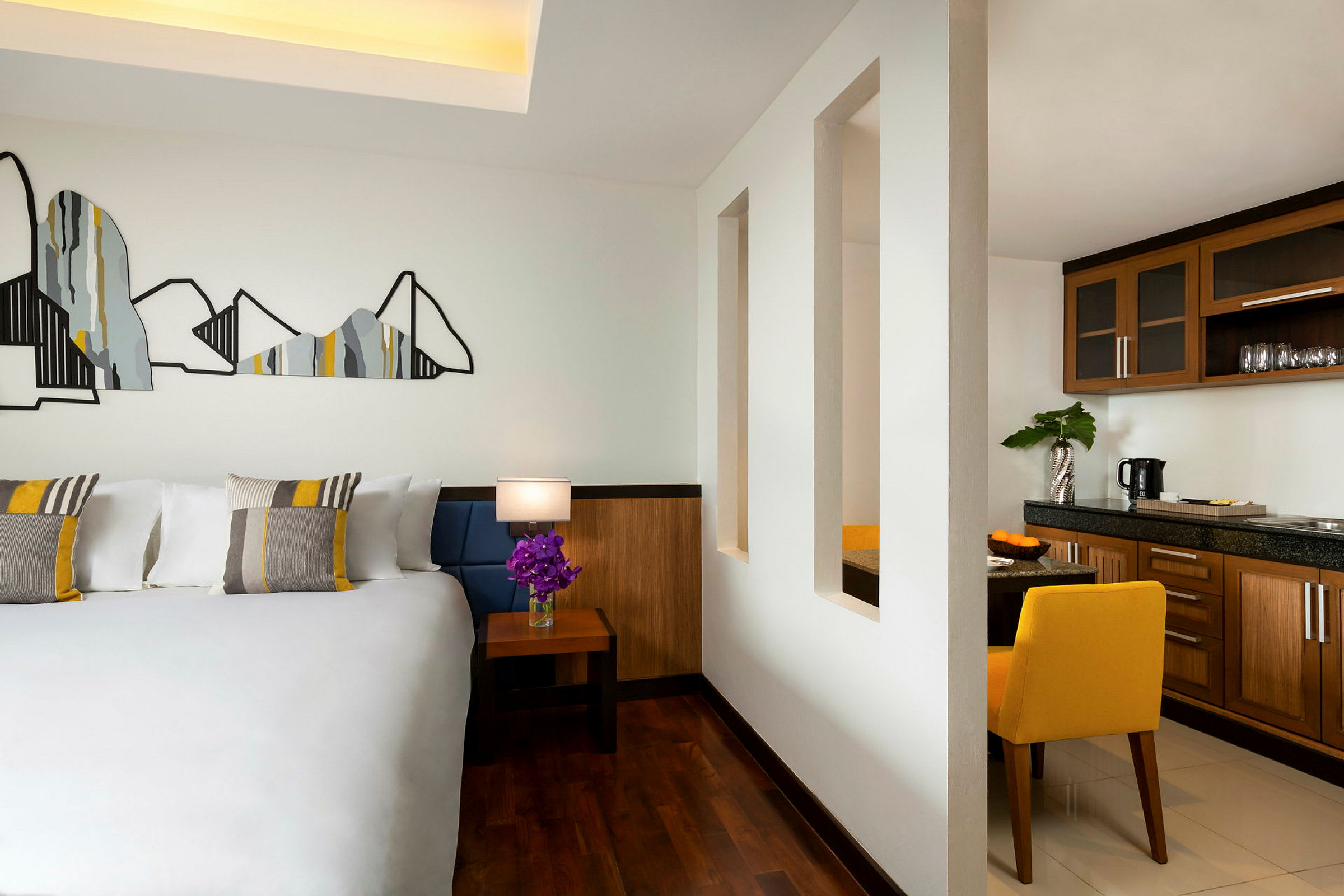 Et hotelværelse med en stor seng med dekorative puder, et sidebord med en lampe og en blomsterbuket, og en åben sektion mod en tekøkken med træindretning. (AI-genereret tekst)