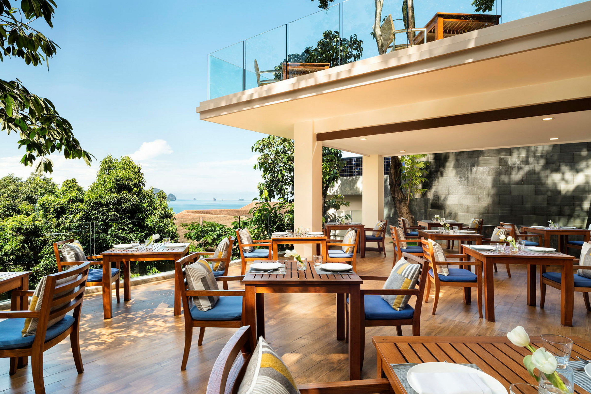 En udendørs restaurant med træborde og stole, omgivet af grønt og med udsigt over havet. (AI-genereret tekst)
