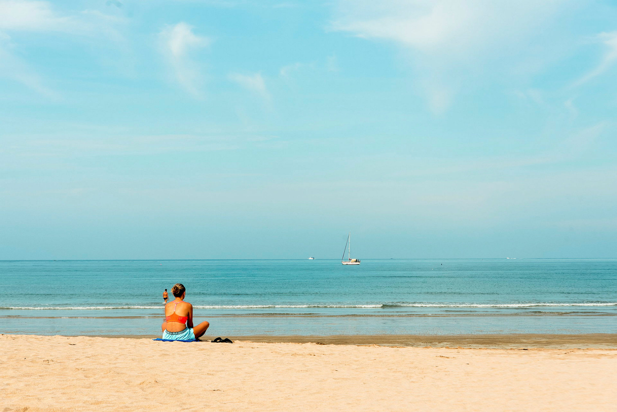En person sidder på en strand og kigger ud over havet, hvor en sejlbåd kan ses i det fjerne. (AI-genereret tekst)