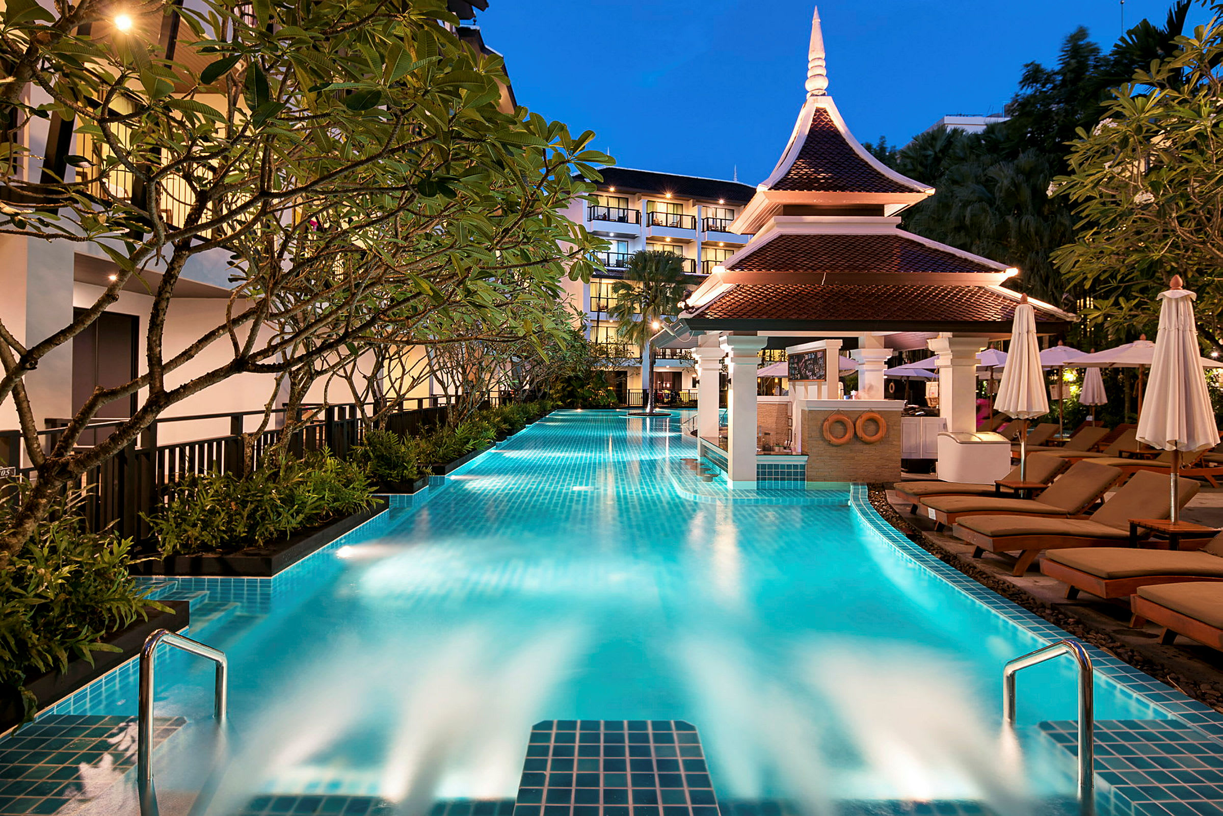 En oplyst pool med træer omkring, liggestole og et tag med moderne thailandsk arkitektur. (AI-genereret tekst)