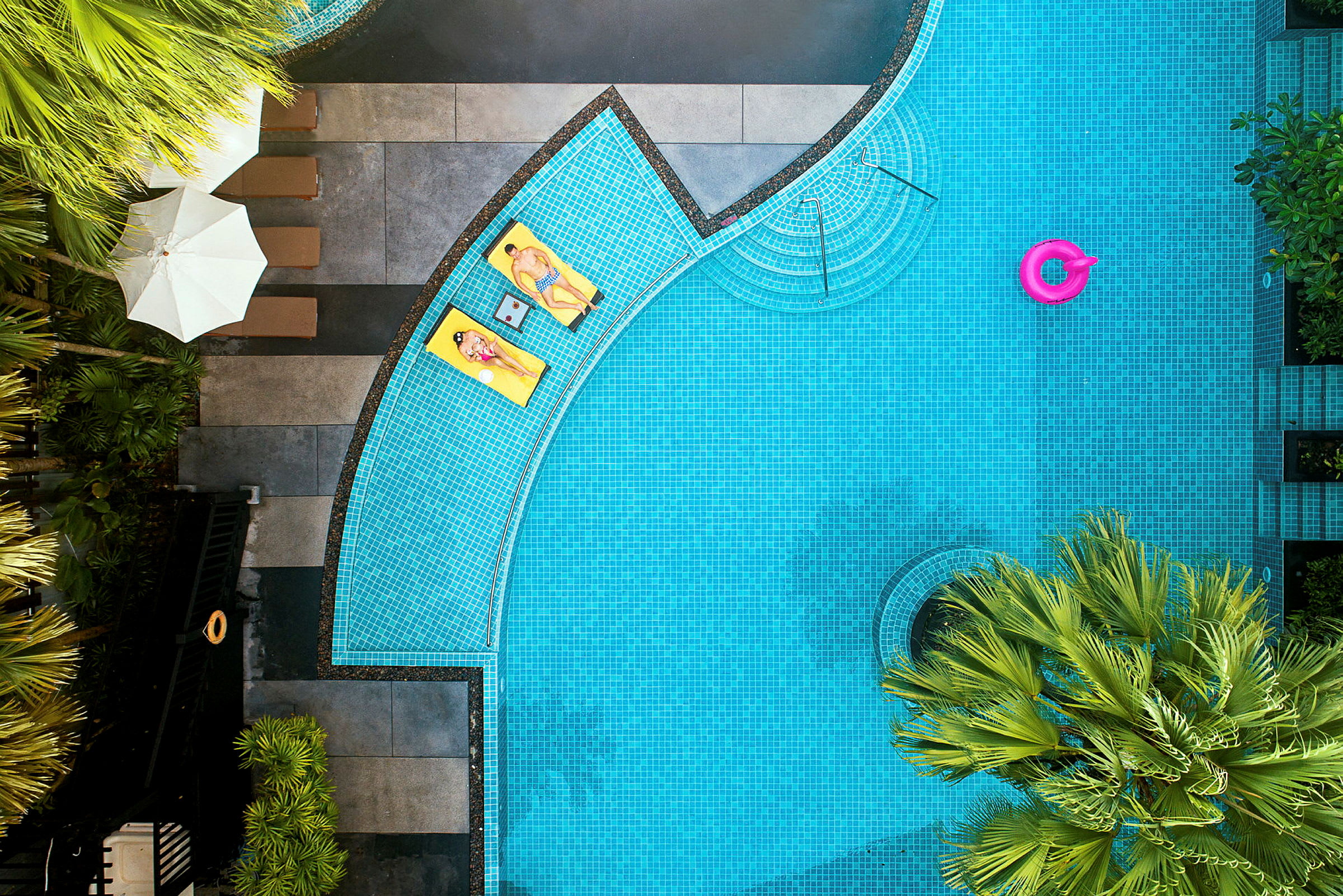 Udsigt ovenfra af en pool med to personer på gule liggestole ved kanten og en pink badering i vandet. Palme og parasoller ses rundt om. (AI-genereret tekst)