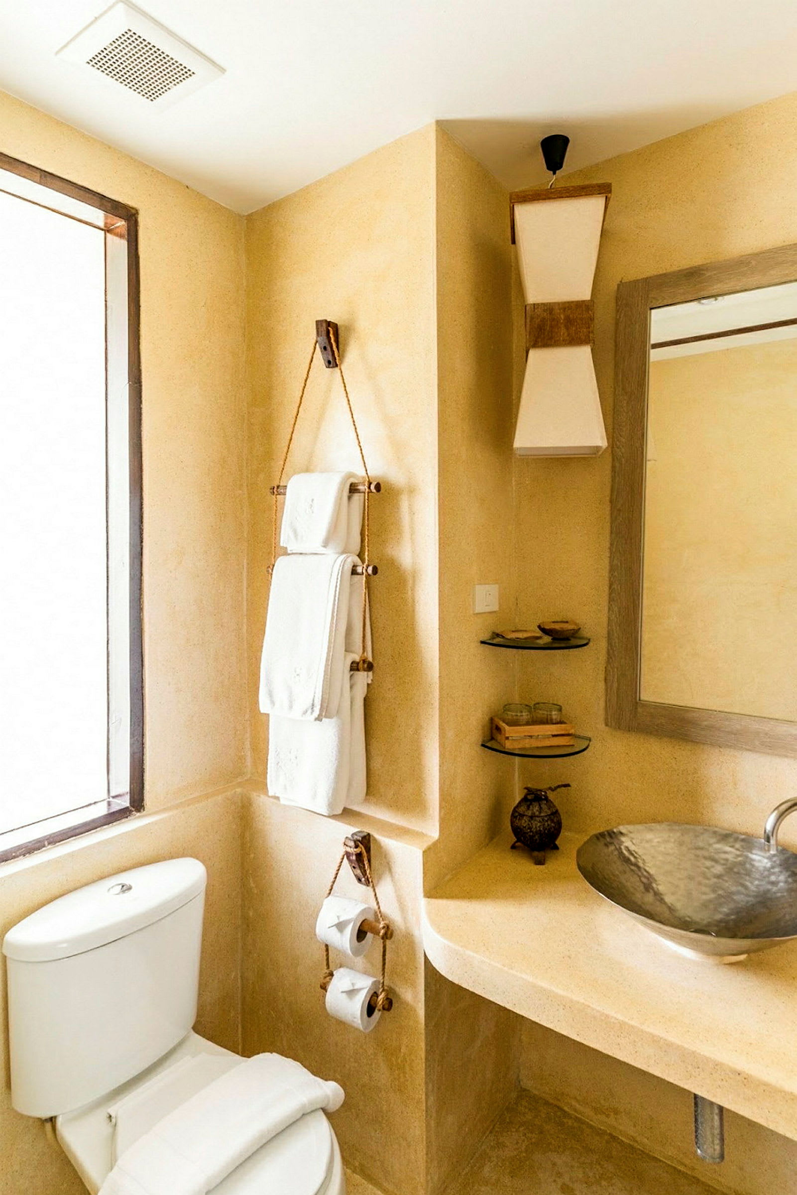 Et badeværelse med beige vægge, et spejl og en håndvask med metallisk overflade. Et toilet er placeret ved siden af med sammenfoldede håndklæder ovenpå. (AI-genereret tekst)