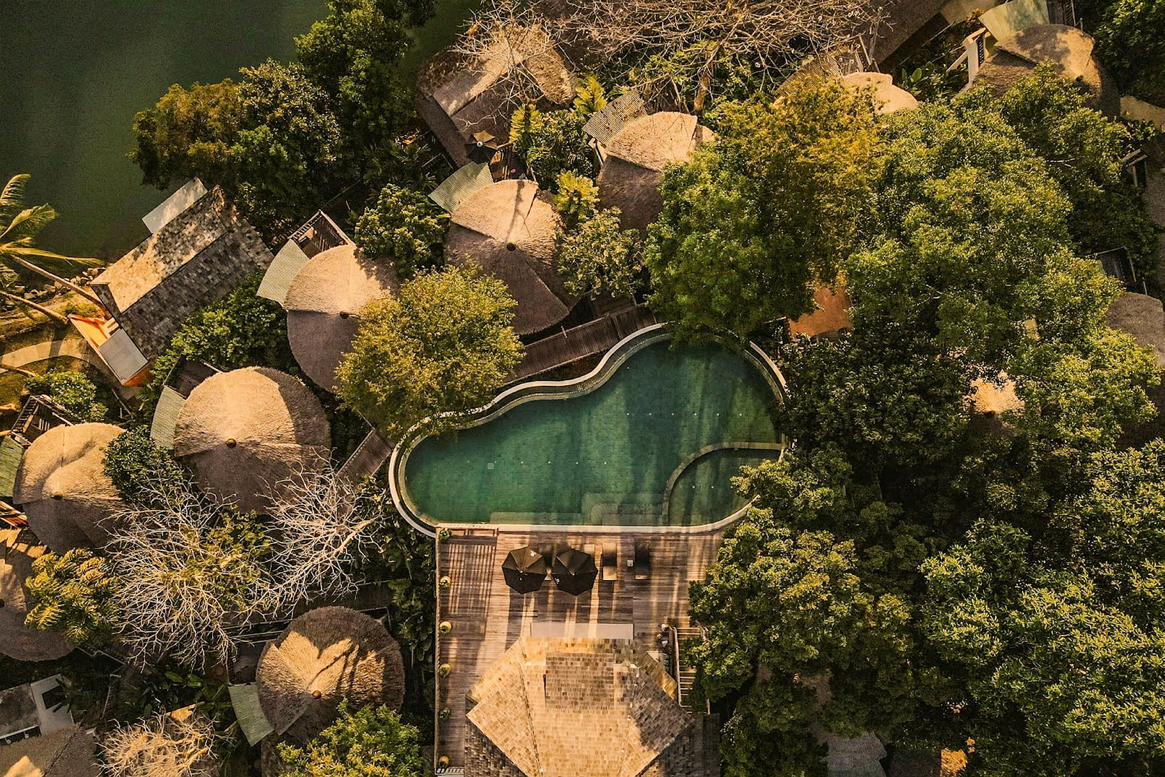 Luftfoto af flere runde huse omgivet af frodig vegetation, ved siden af en uregelmæssig pool og en træterrasse. (AI-genereret tekst)