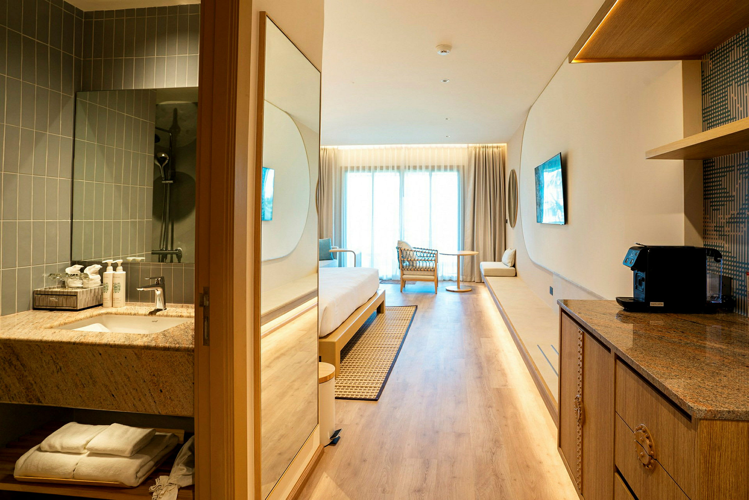 Et moderne hotelværelse med lyst interiør, herunder seng, lænestol, lille bord og altan. Et badeværelse og et køkkenområde ses i forgrunden. (AI-genereret tekst)