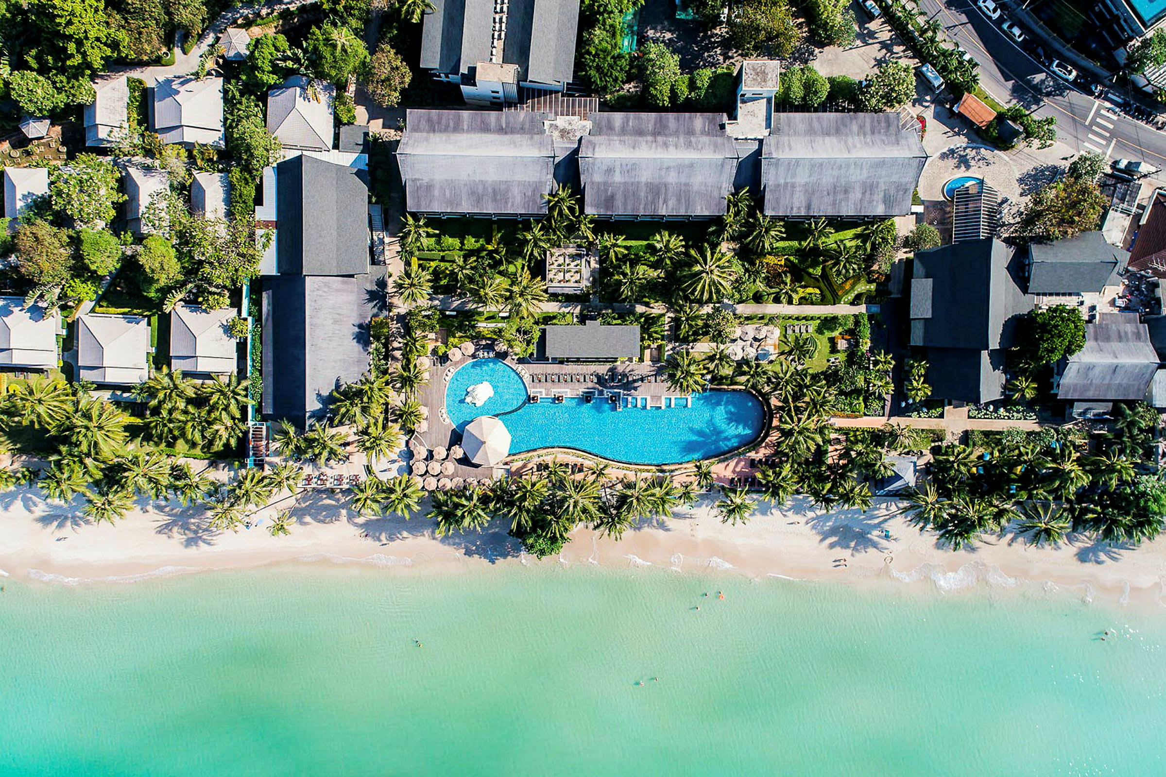 Luftfoto af en ferieresort med pool, omgivet af tropiske træer og tæt på en strand med klart vand. (AI-genereret tekst)