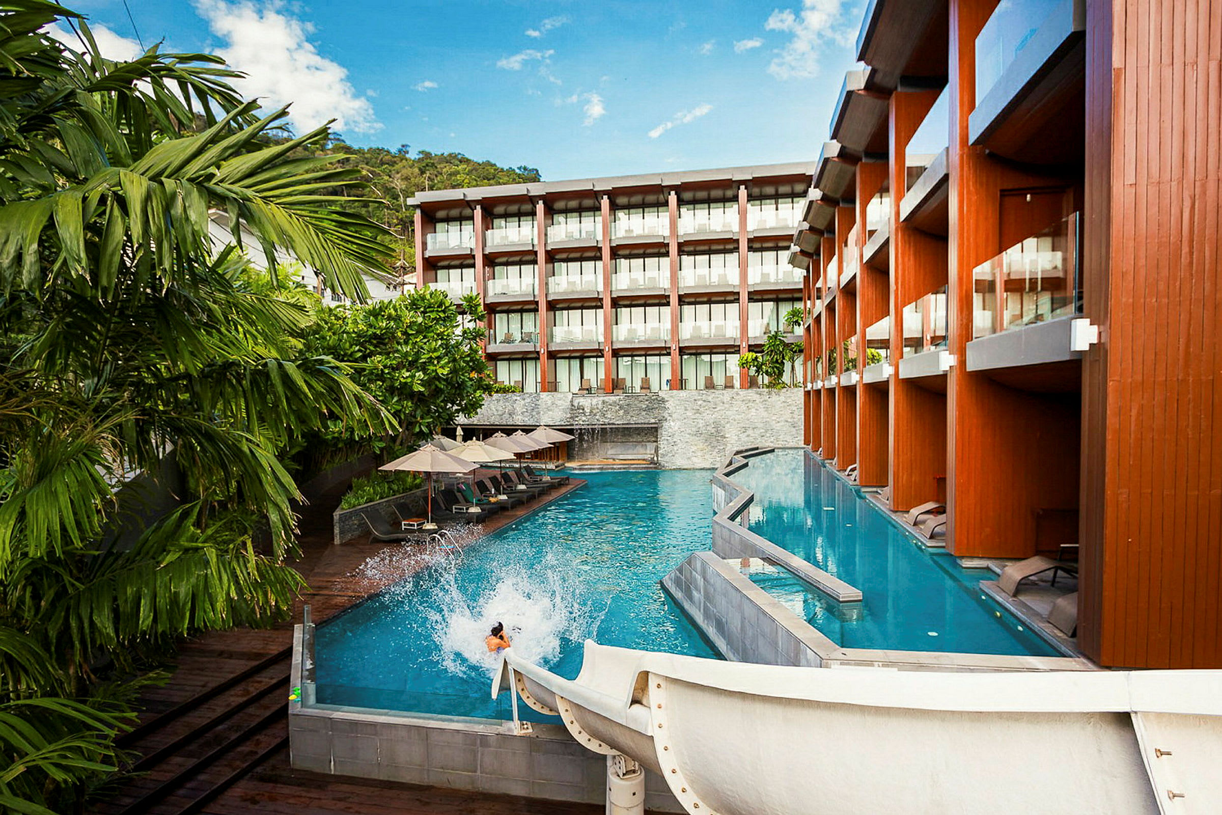 En pool ved et moderne hotel med liggestole og parasoller samt en vandrutsjebane, der fører ned i vandet. (AI-genereret tekst)