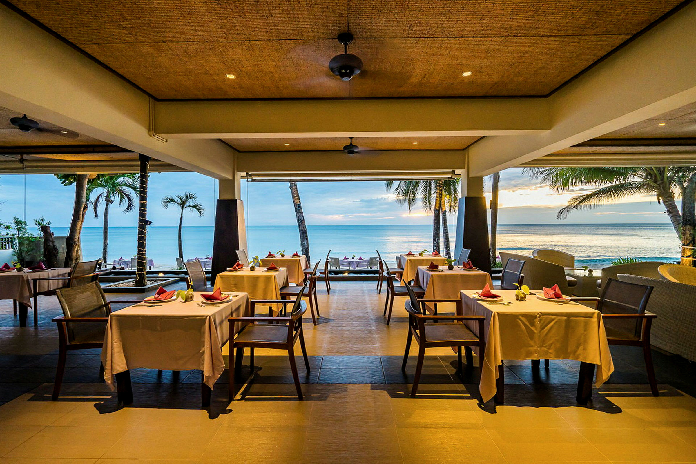 En restaurant ved havet med hvide duge på bordene og udsigt over palmer og stranden. (AI-genereret tekst)