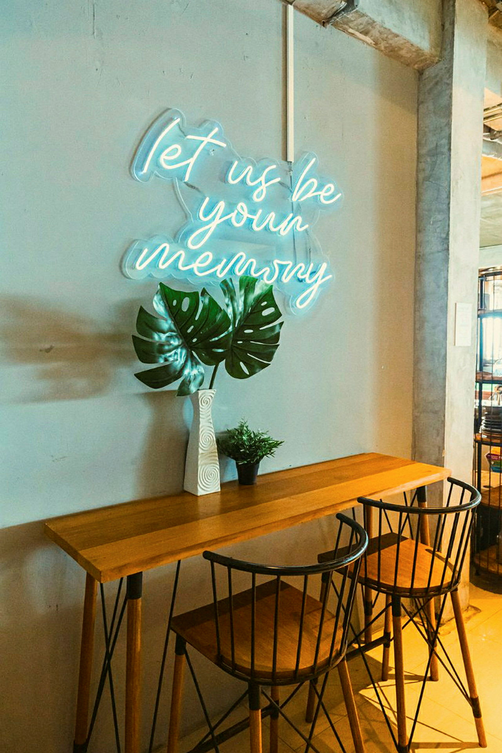 Et bord med to stole, en vase med store grønne blade og en lille grøn plante, foran en væg med et neonskilt med teksten 'let us be your memory'. (AI-genereret tekst)
