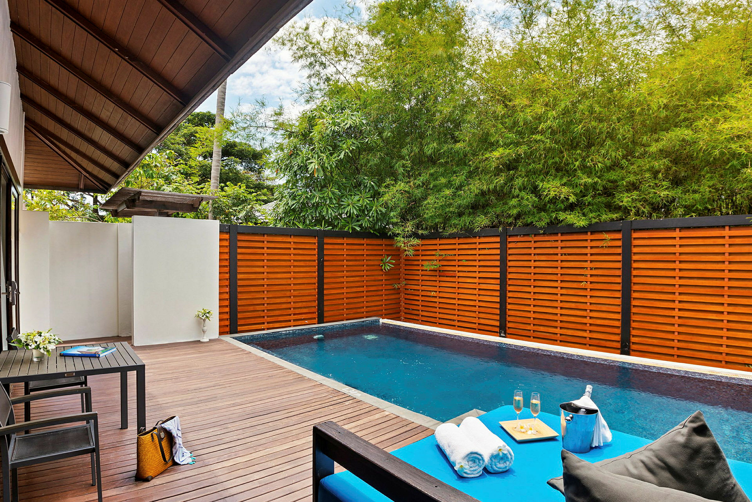 En privat pool med træterrasse, omgivet af træhegn og grøn beplantning. En lounge med håndklæder, drikkevarer og en serveringsbakke ses i forgrunden. (AI-genereret tekst)