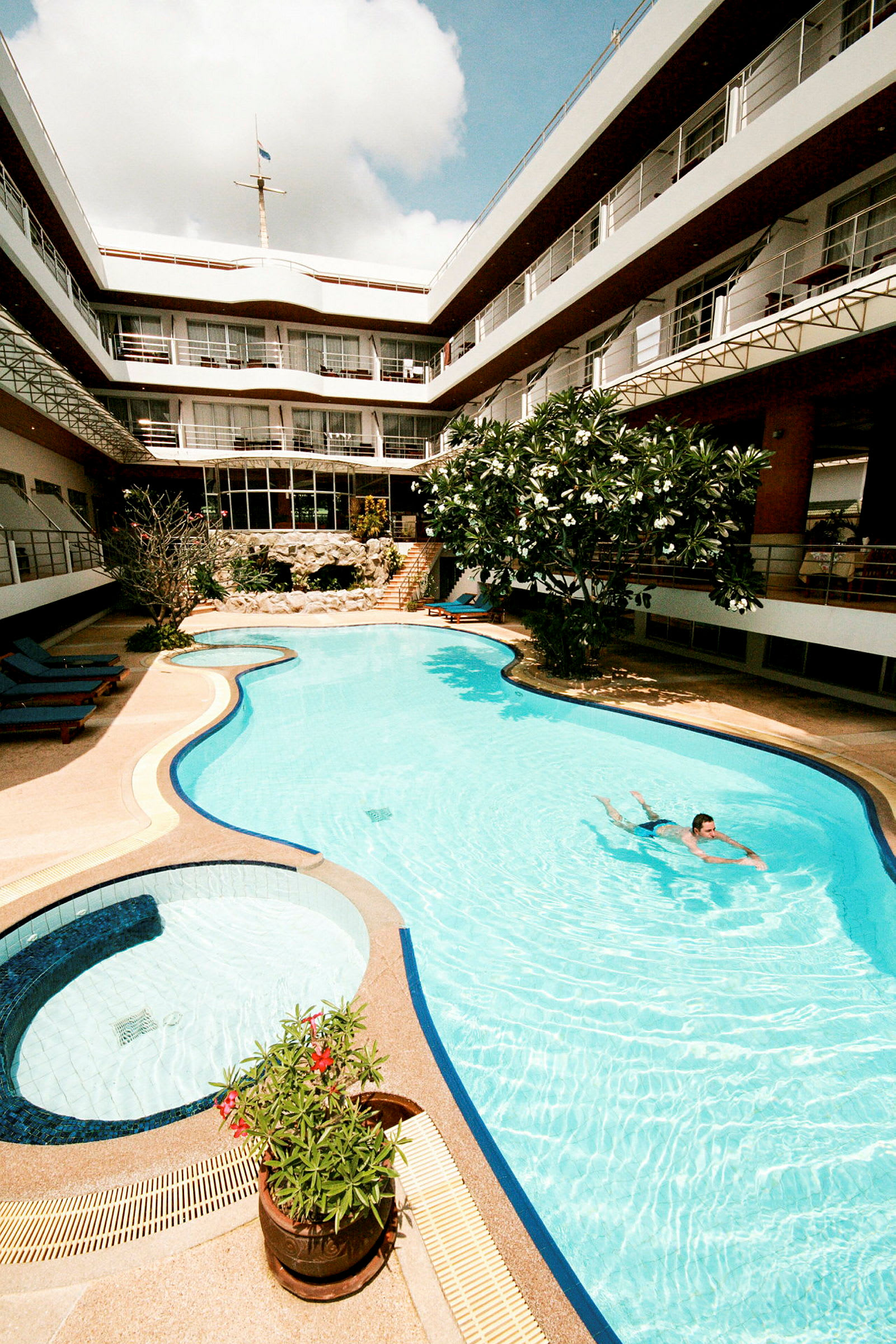 En pool omgivet af en bygning med flere etager og altaner. Der er liggestole og grønne planter omkring poolen. (AI-genereret tekst)