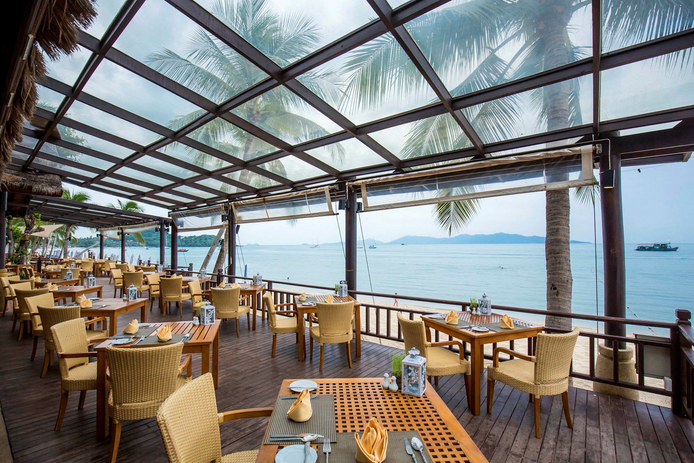 En restaurant med udendørs servering under et glastag. Stole og borde er placeret på en træterrasse med udsigt over stranden og havet. (AI-genereret tekst)