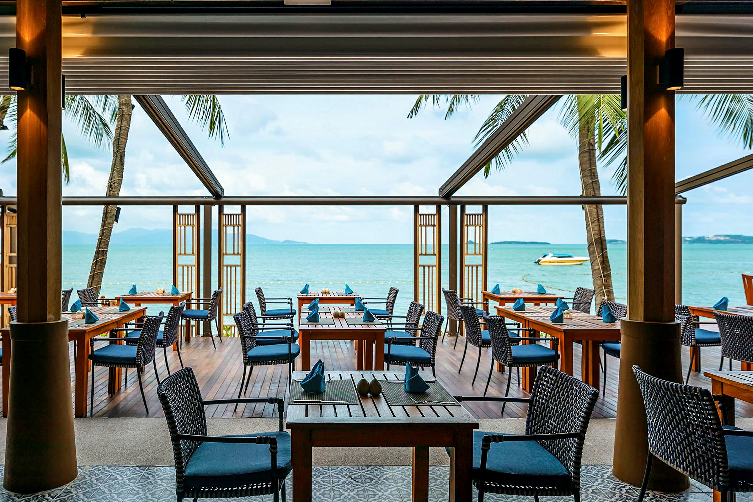 En udendørsrestaurant med træborde og stole ligger ved havet og tilbyder udsigt over vandet og en båd. (AI-genereret tekst)