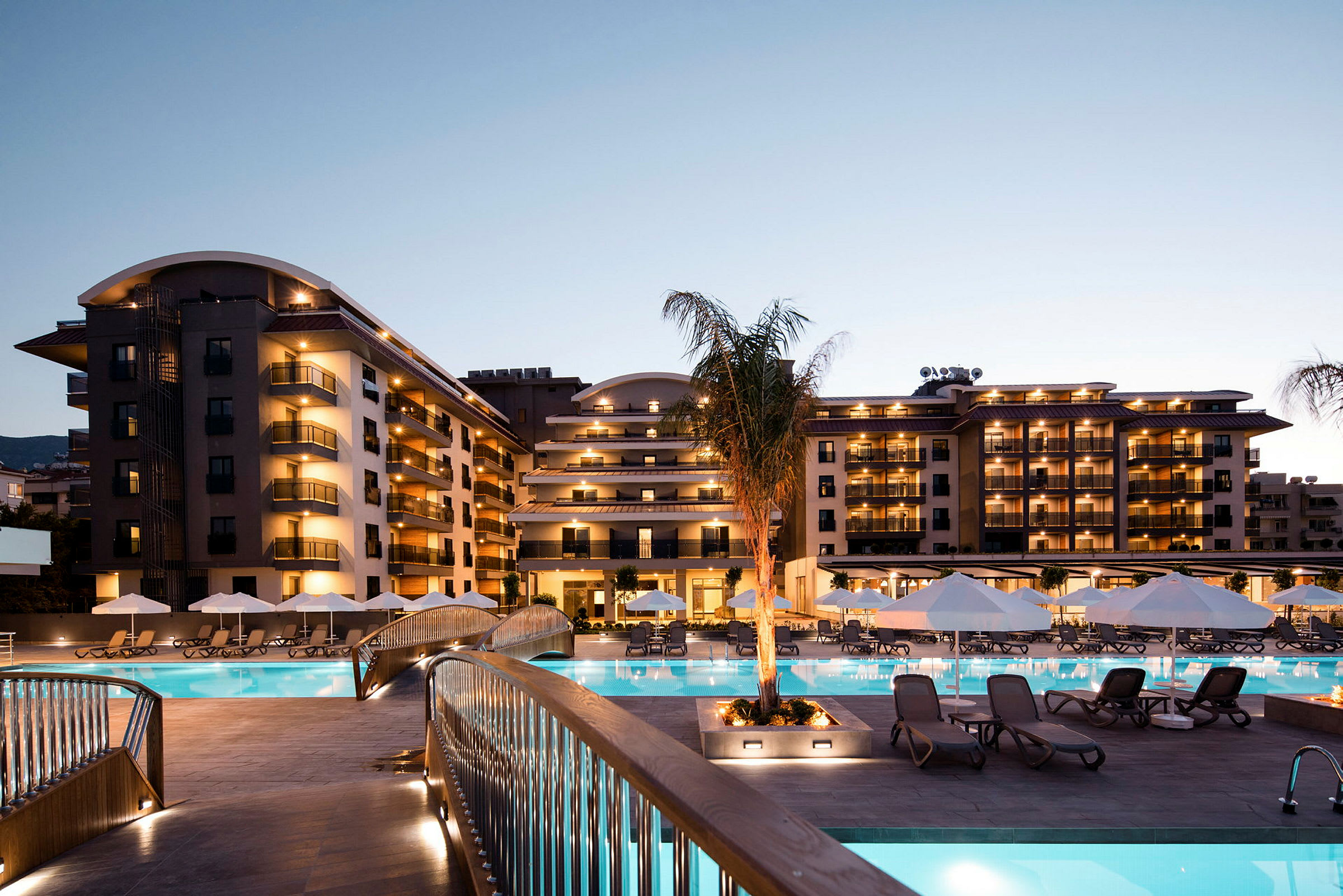 Moderne hotelbygning ved udendørs pool med solsenge og parasoller, oplyst om aftenen. (AI-genereret tekst)