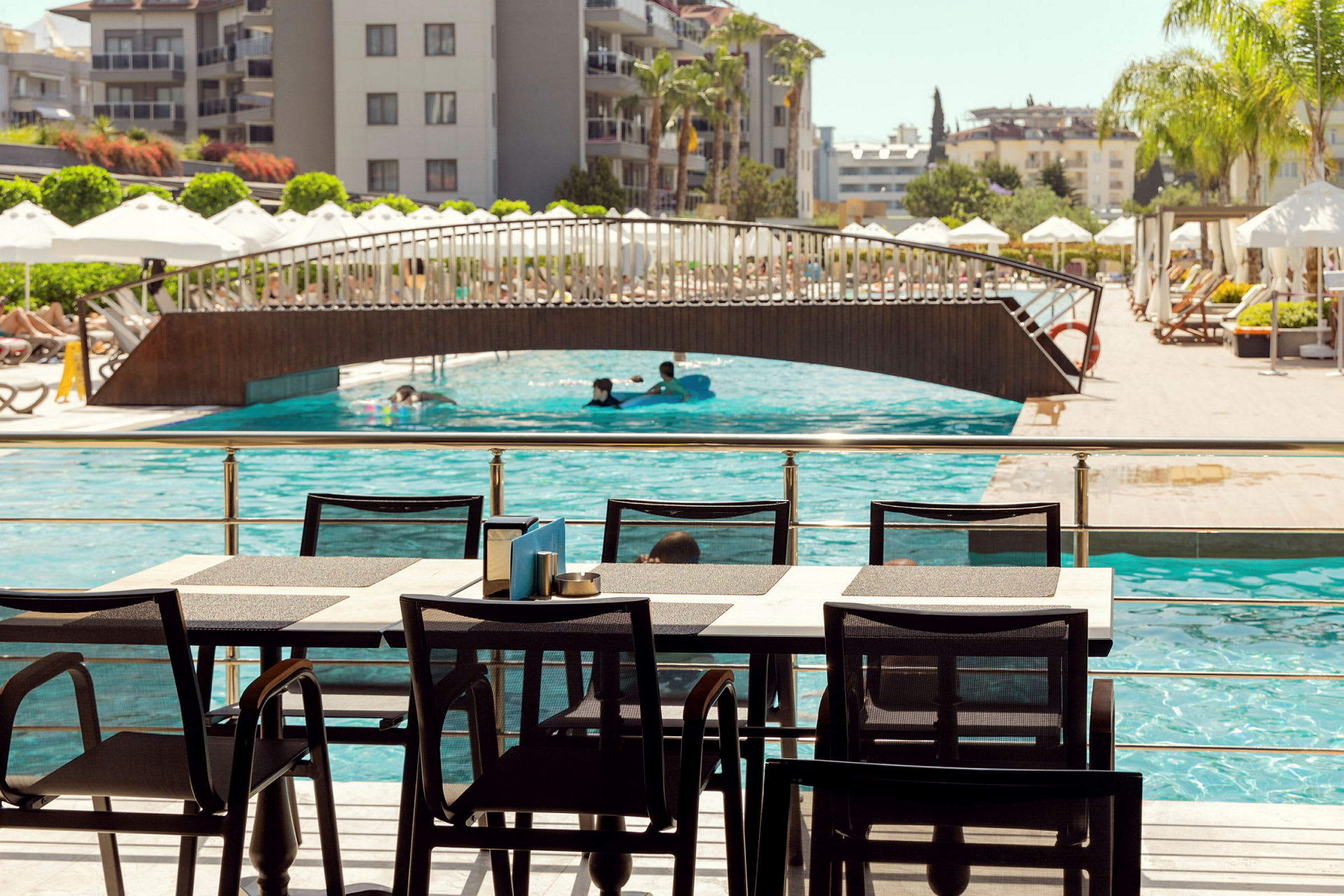 Spiseområde ved en pool med bro og hotel i baggrunden. (AI-genereret tekst)