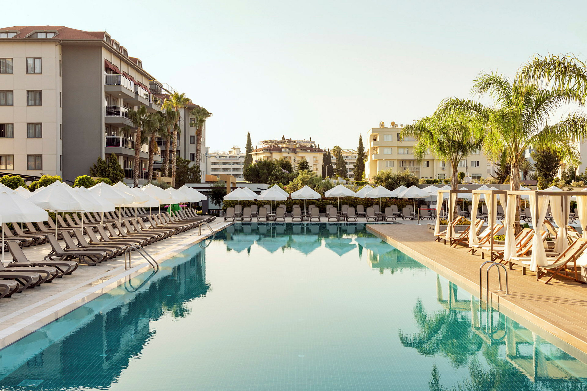 En hotelpool med liggestole og parasoller ved siden, omgivet af bygninger og palmer. (AI-genereret tekst)